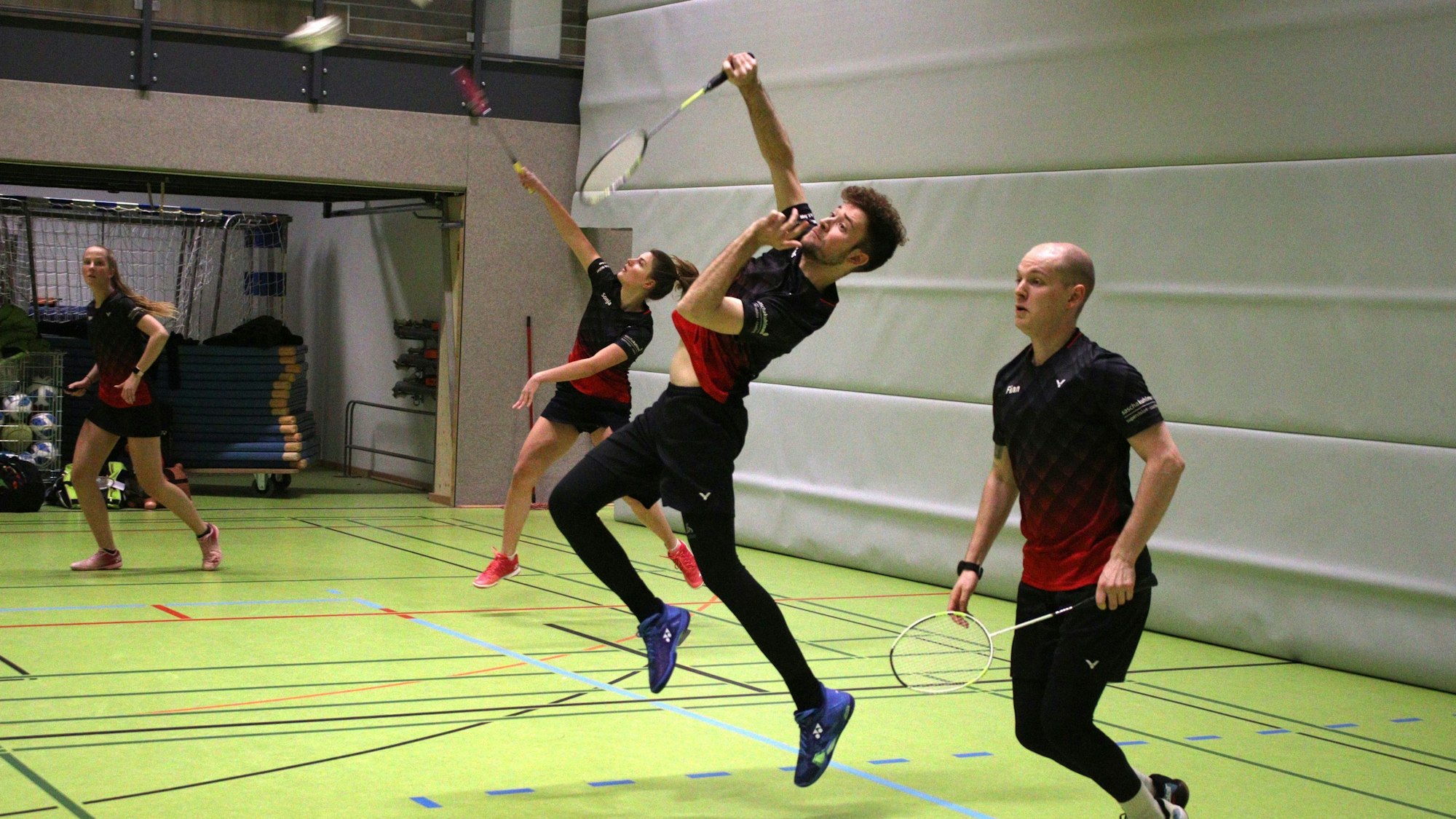 Vier Kaller Badmintonspieler bilden zwei Doppelpaare und spielen ihre Partien. Im Hintergrund ist ein offener Abstellraum zu sehen.