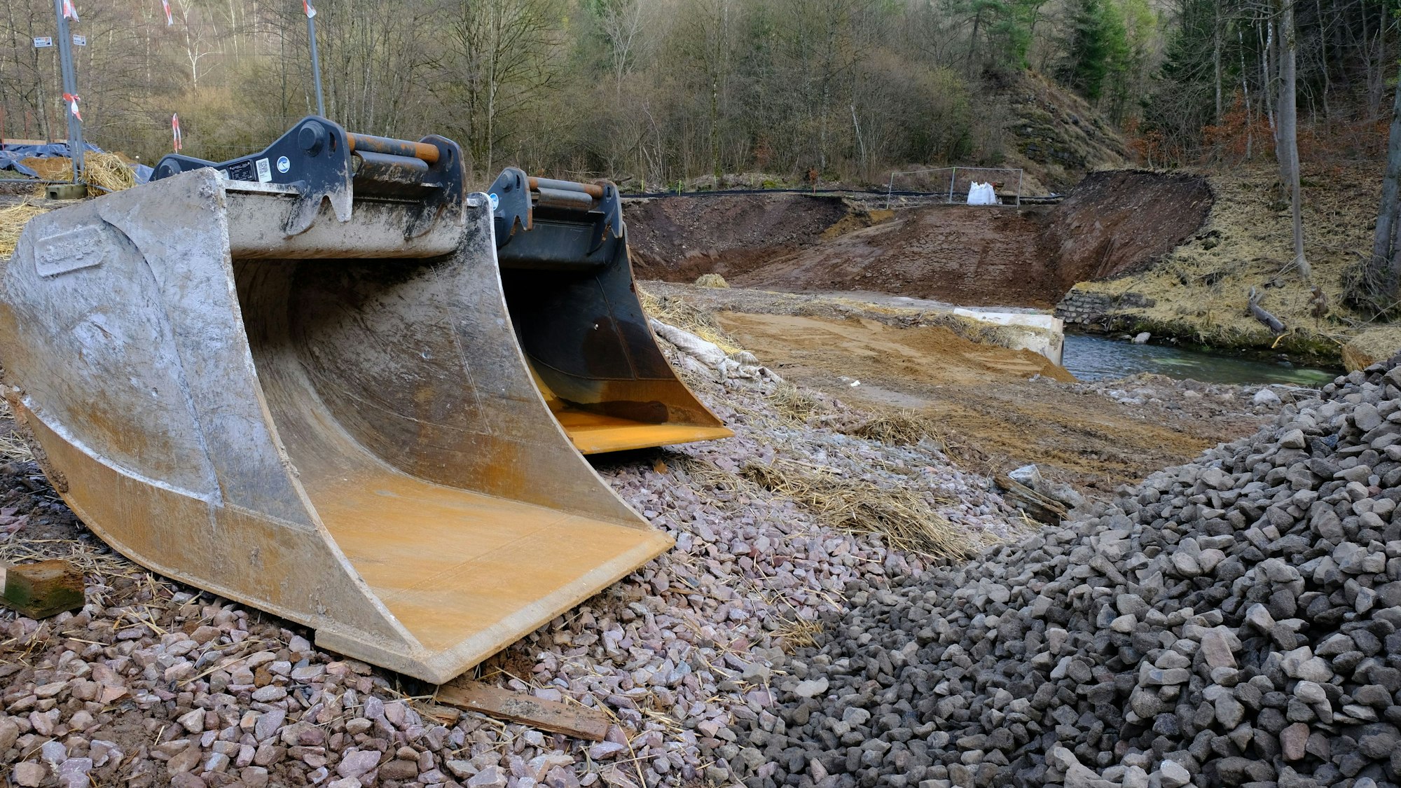 An einer Baustelle liegen zwei große Baggerschaufeln. Im Hintergrund ist das Flusstal der Urft zu erkennen.