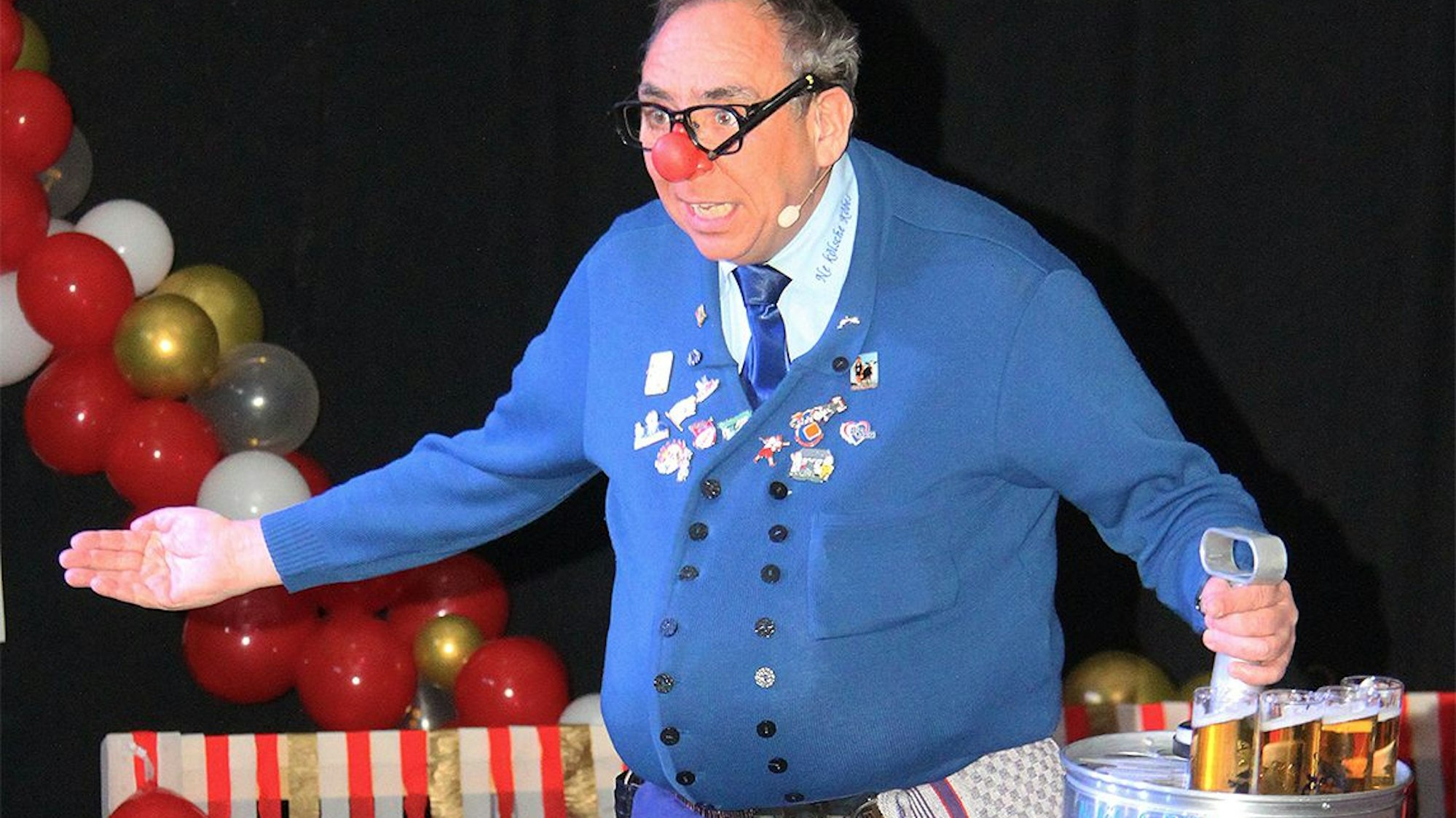 Das Bild zeigt Axel Höfel auf der Bühne. In seiner linken Hand hält einen Kranz mit Kölsch.