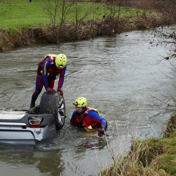 Zwei Männer in Neoprenanzügen stehen in einem Fluss an einem Auto, bei dem nur die Hinterräder aus dem Wasser ragen.