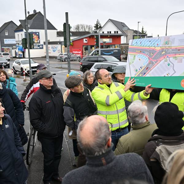 Ein Mitarbeiter des Planungsbüros in gelber Weste hält einen farbigen Entwurf, der die Gestaltung der Kreuzung zeigt, hoch. Teilnehmer der Veranstaltung stehen drumherum.