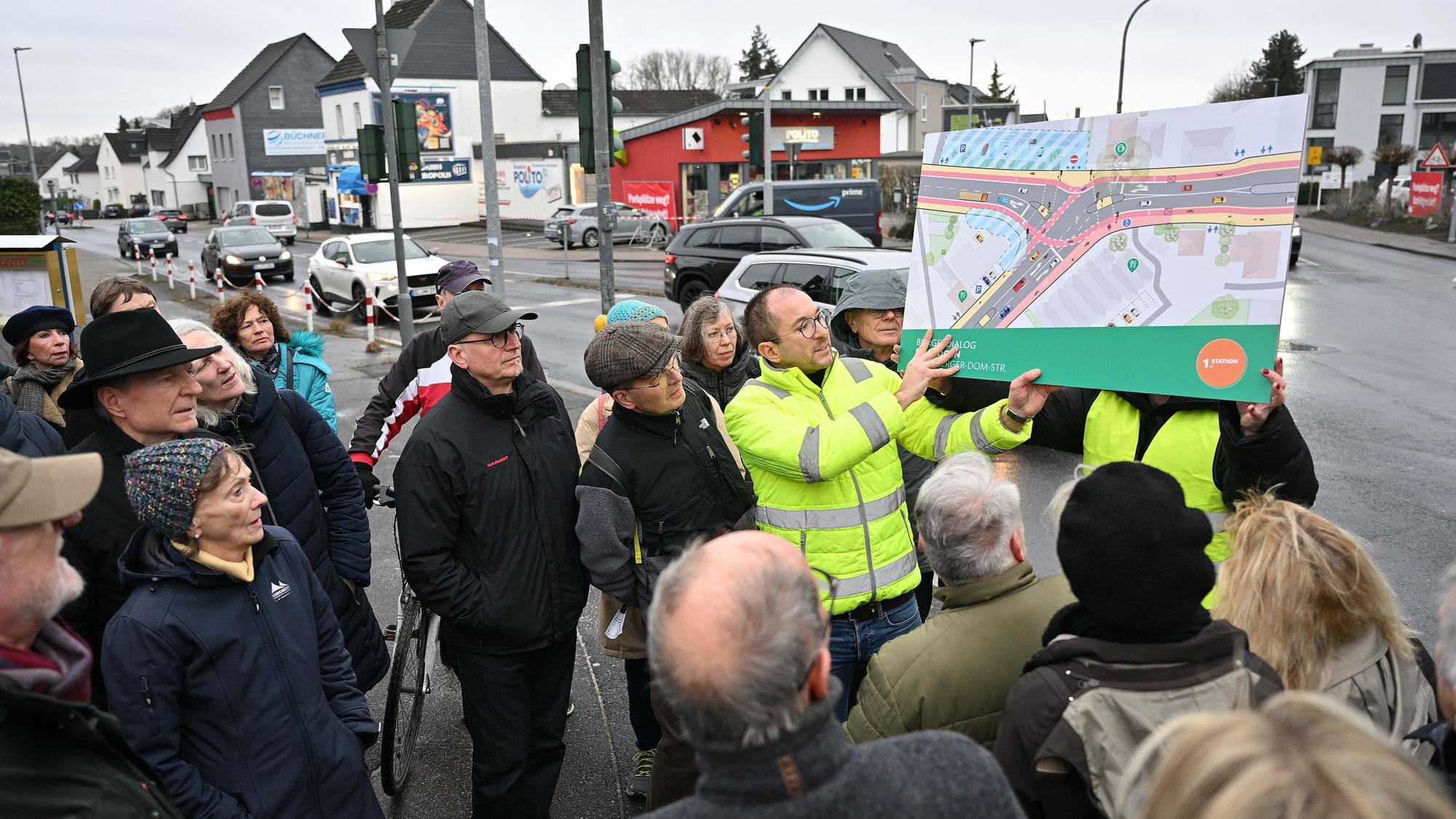 Zwei Planer halten ein Plakat hoch, das die vorgesehene Verkehrsführung zeigt. Um sie herum stehen Teilnehmer der Veranstaltung. Im Hintergrund sieht man die Straßenkreuzung.