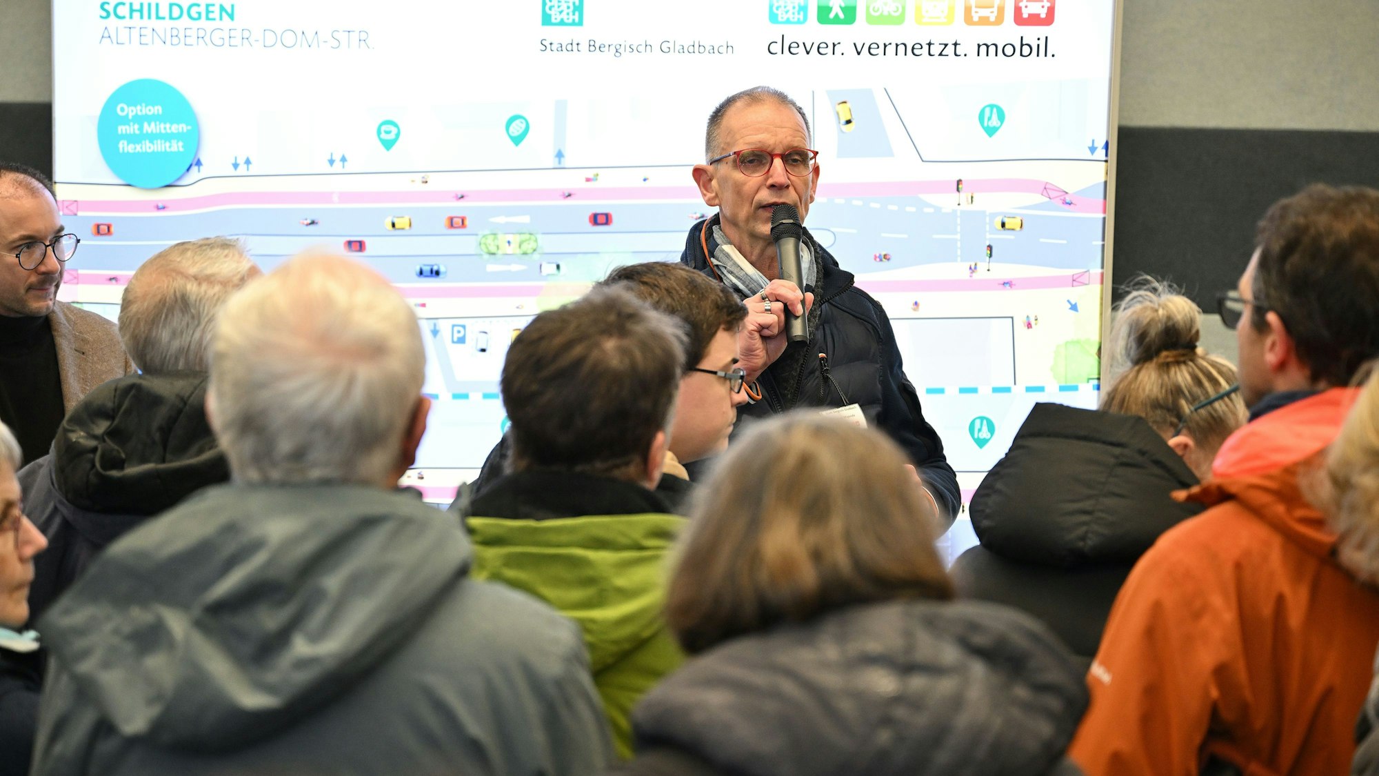 Ragnar Migenda spricht in ein Mikrophon. Dahinter ist die Tafel mit der Ansicht der Planung zu sehen. Im Vordergrund sieht man Zuhörer.