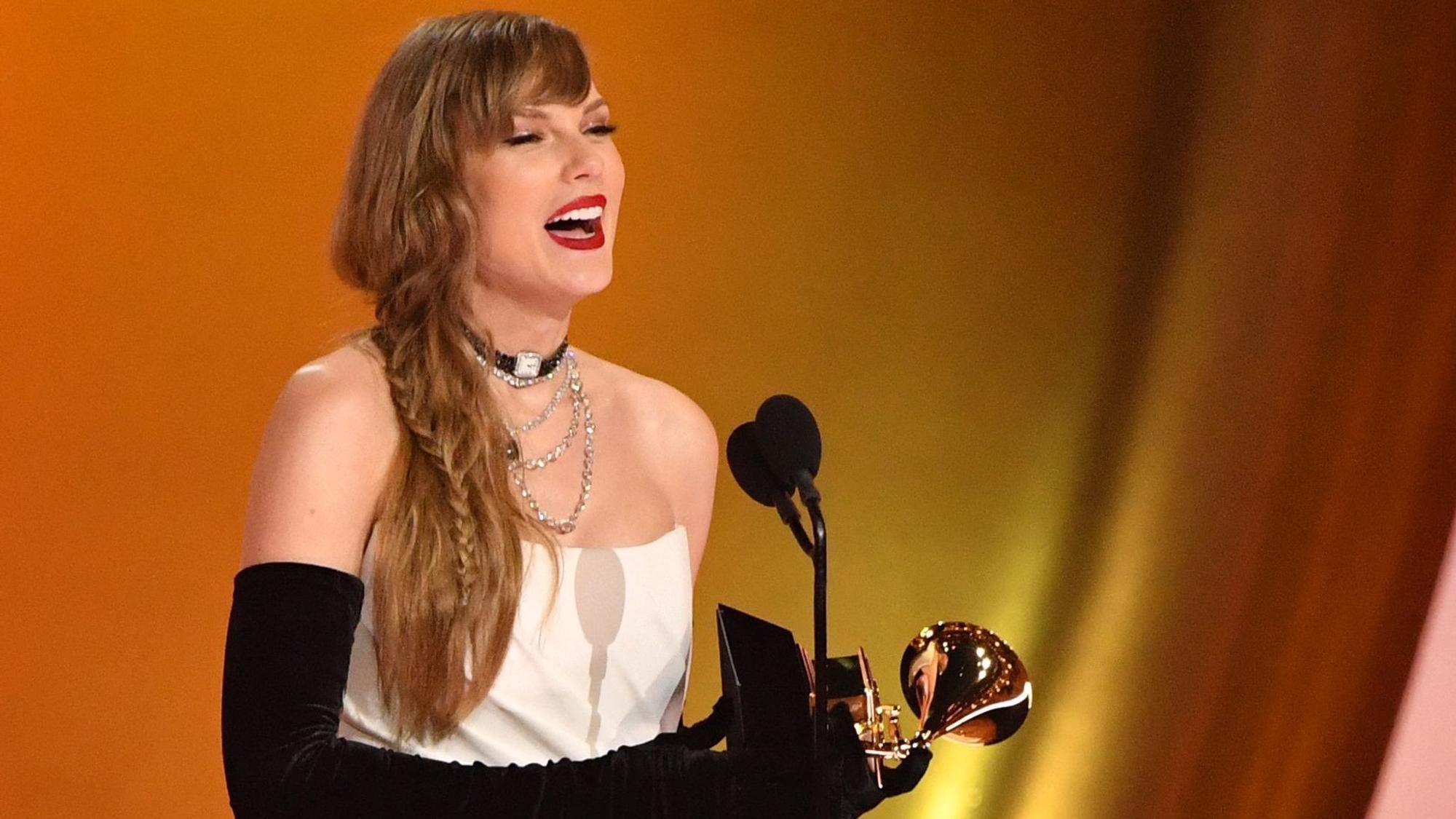 Taylor Swift hält einen Grammy-Preis in der Hand. Sie trägt ein weißes schulterloses Kleid und lange schwarze Handschuhe.