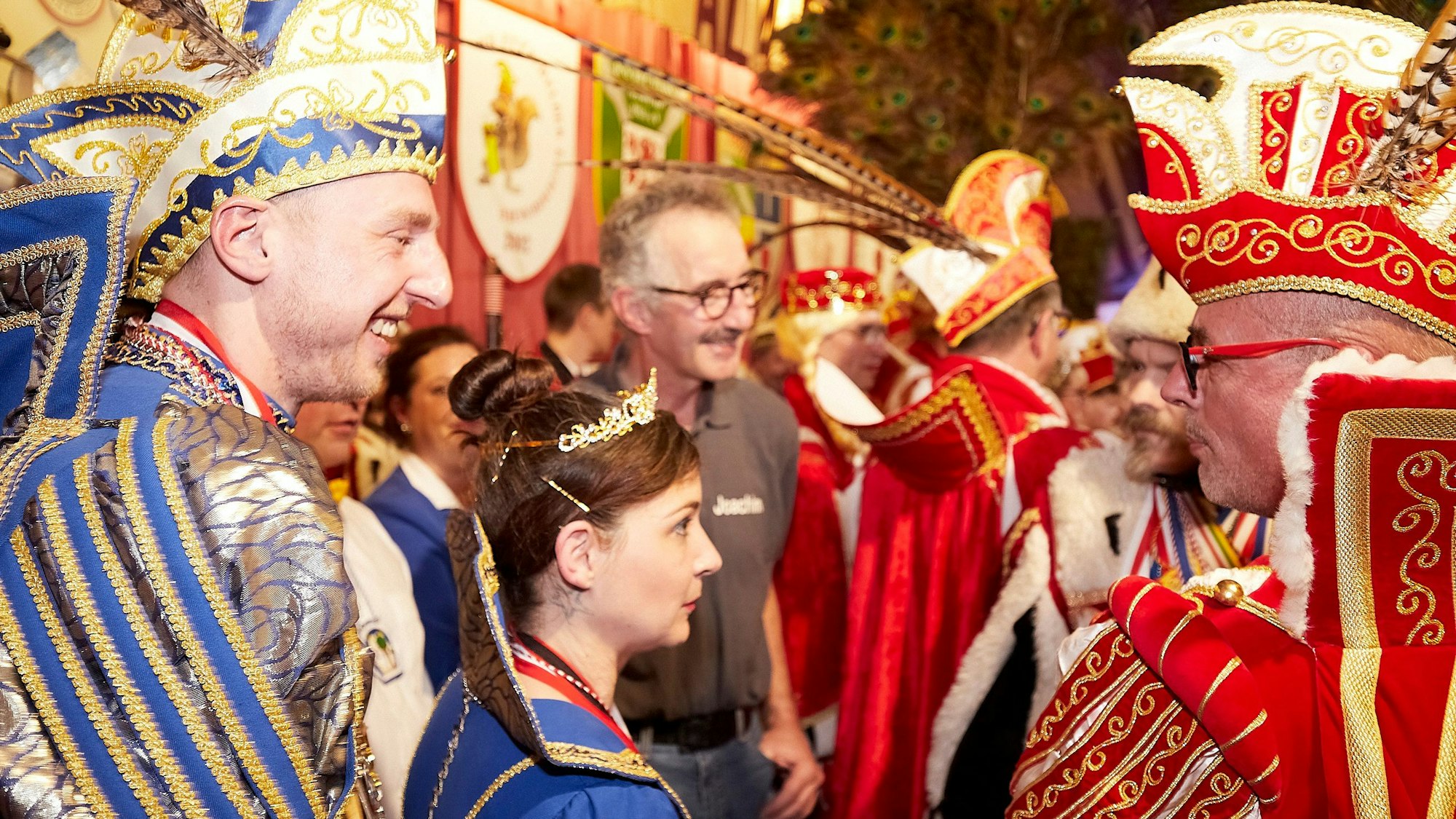 Zahlreiche Karnevals-Tollitäten stehen auf einer Bühne. Ein Prinz in rot-weißem Kostüm unterhält sich mit einem Prinzenpaar in blau-weißem Ornat.