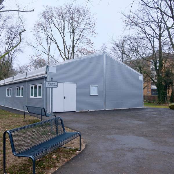 Weil die alte Trauerhalle auf dem Melatenfriedhof seit neun Monaten saniert werden soll, finden die den Trauerfeiern in einer grauen Leichtbauhalle statt, die an eine Verrichtunsgbox erinnert.
