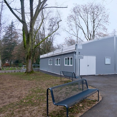 Weil die alte Trauerhalle auf dem Melatenfriedhof seit neun Monaten saniert werden soll, finden die den Trauerfeiern in einer grauen Leichtbauhalle statt, die an eine Verrichtunsgbox erinnert.