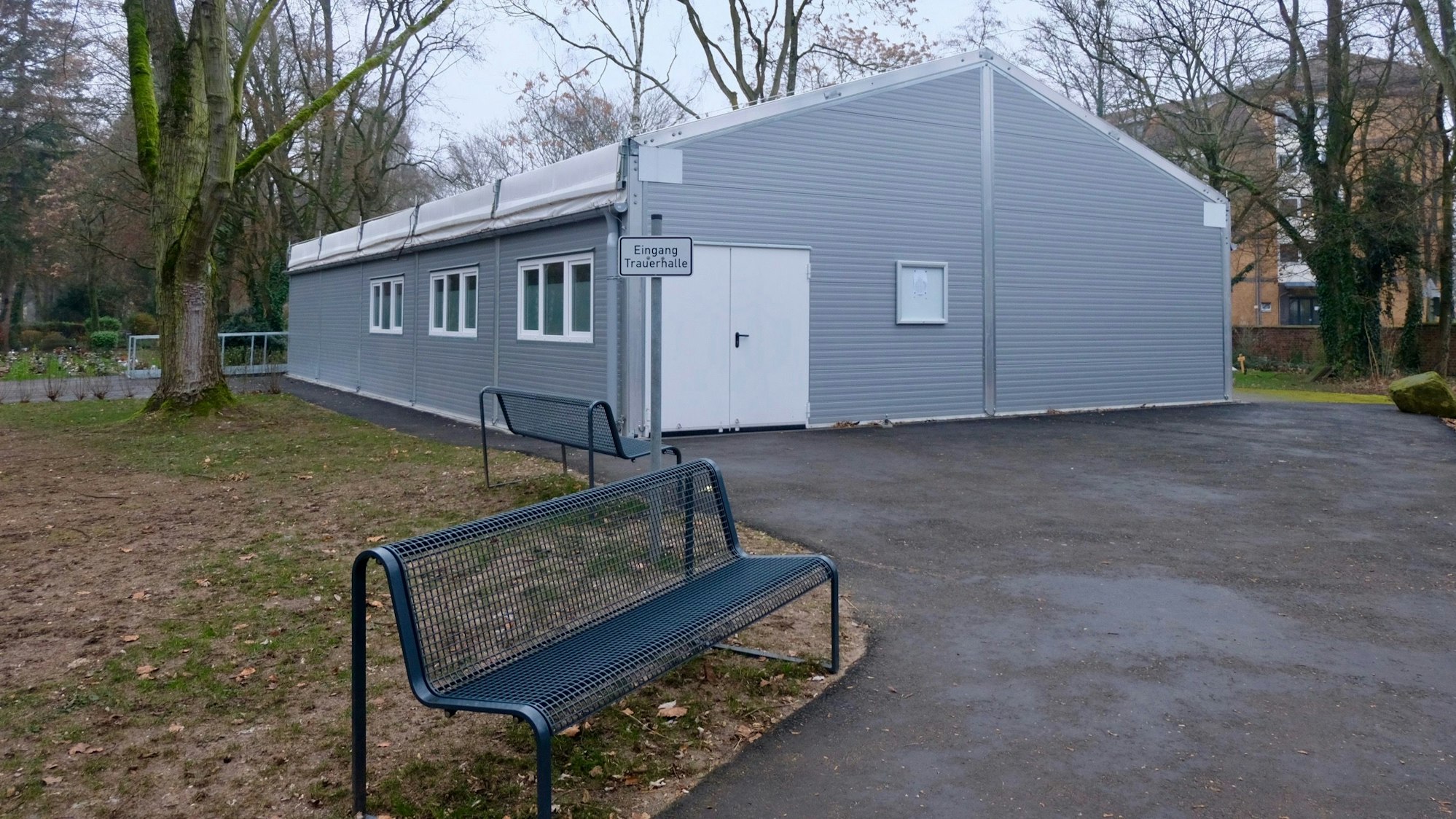 Weil die alte Trauerhalle auf dem Melatenfriedhof seit neun Monaten saniert werden soll, finden die den Trauerfeiern in einer grauen Leichtbauhalle statt, die an eine Verrichtunsgbox erinnert.