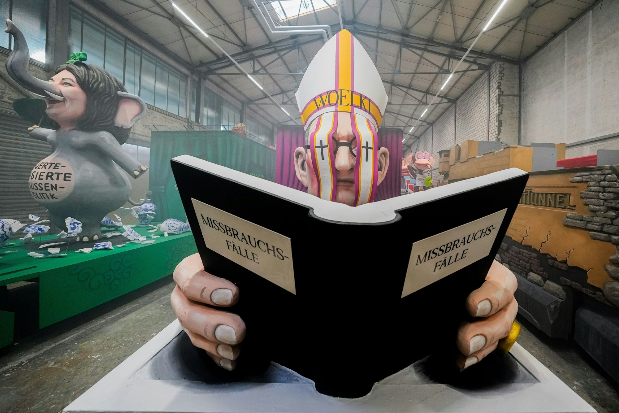 Ein Motivwagen, der Kardinal Woelki mit einem Buch mit der Aufschrift ·Missbrauchsfälle· zeigt, steht beim Richtfest für den Kölner Rosenmontagszug im Karnevalsmuseum.