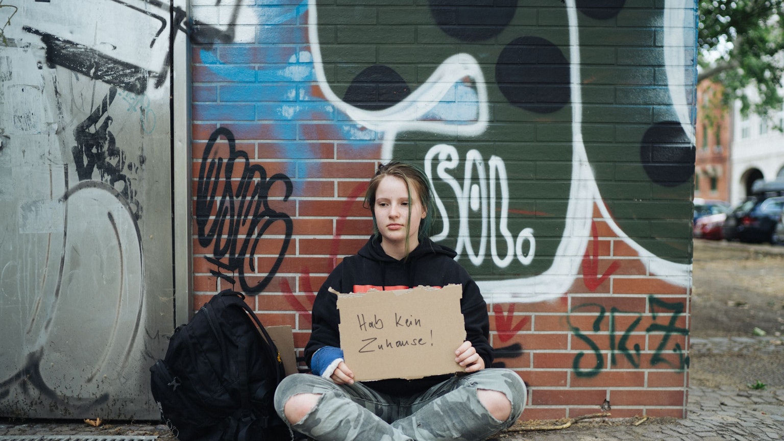 Ein junges obdachloses Mädchen sitzt vor einer Graffiti-Wand und hält ein Pappschild mit der Aufschrift„ Ich habe kein Zuhause“ in der Hand.