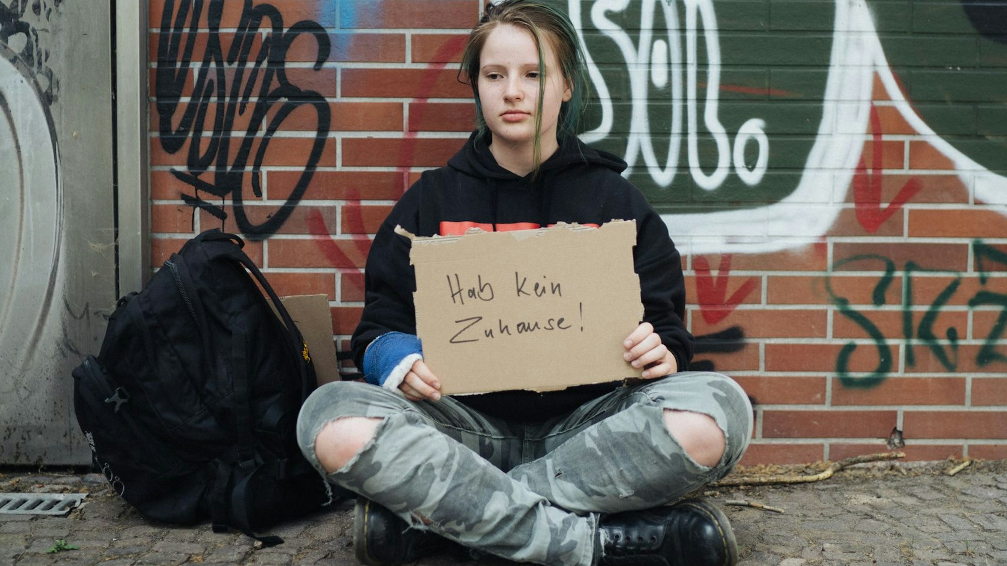 Ein junges obdachloses Mädchen sitzt vor einer Graffiti-Wand und hält ein Pappschild mit der Aufschrift„ Ich habe kein Zuhause“ in der Hand.