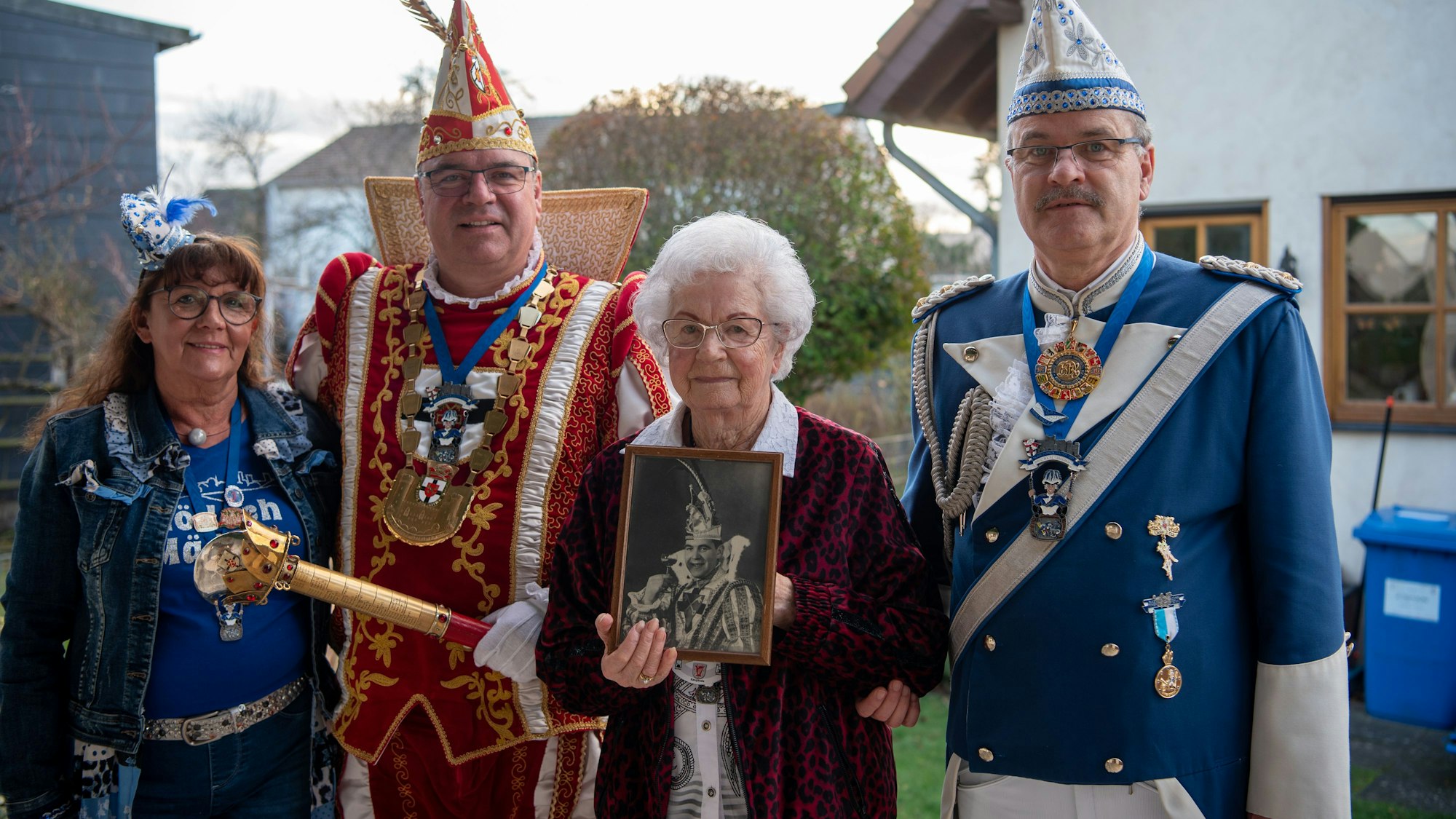 Elisabeth Esser steht auf ihrer Terrasse und hält ein Bild ihres Vaters als Karnevalsprinz in den Händen. Links und rechts neben ihr stehen ihre beiden Söhne Ralf und Udo. Ralf Esser trägt das Ornat des Prinzen und Udo Esser die Uniform der Blauen Funken. Neben Ralf Esser steht seine Frau Anne-Marie.