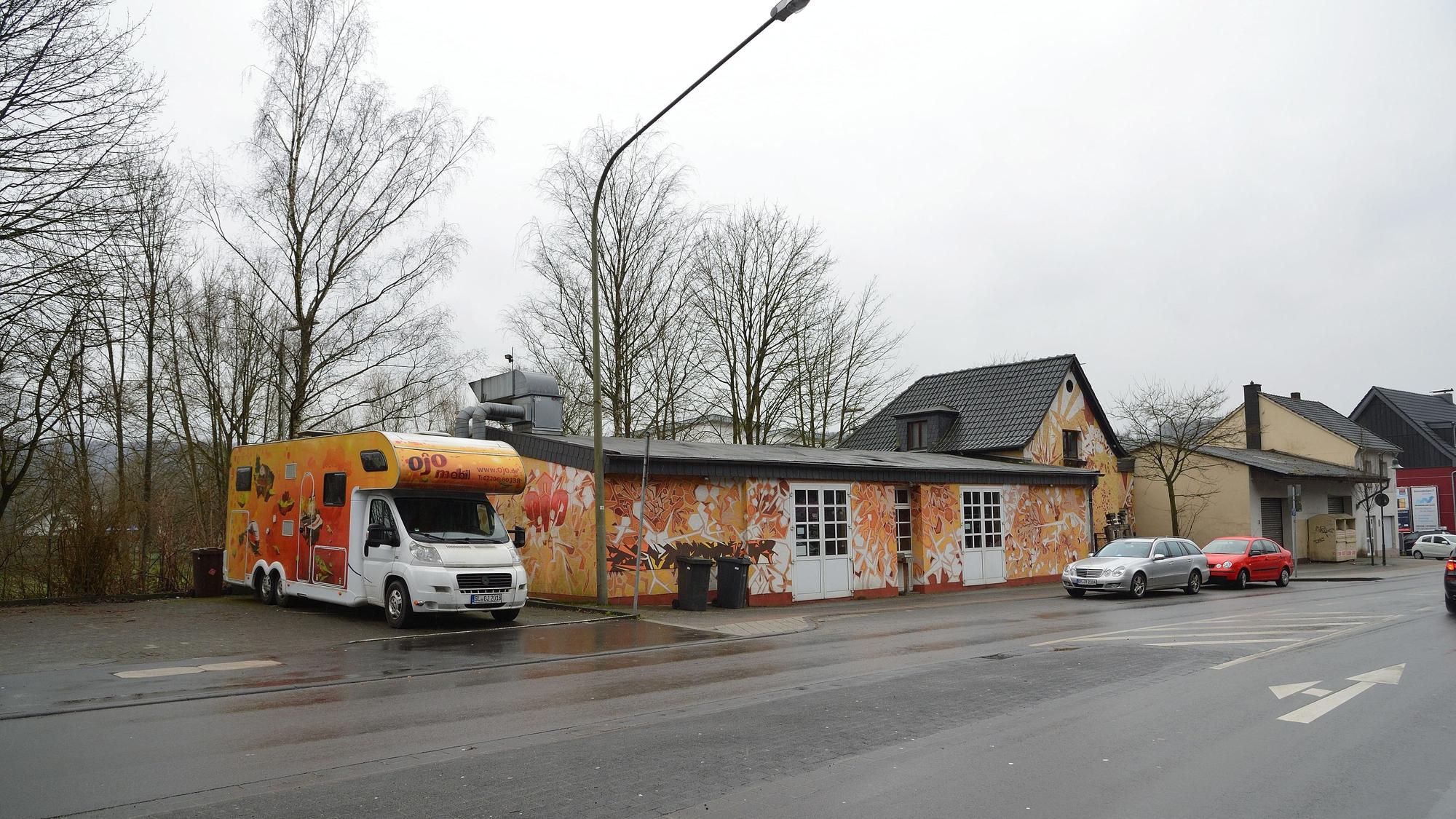 Ein gelb- und orangefarben angemaltes Haus steht an einer Hauptstraße, daneben befindet sich ein Wohnmobil in den selben Farben.
