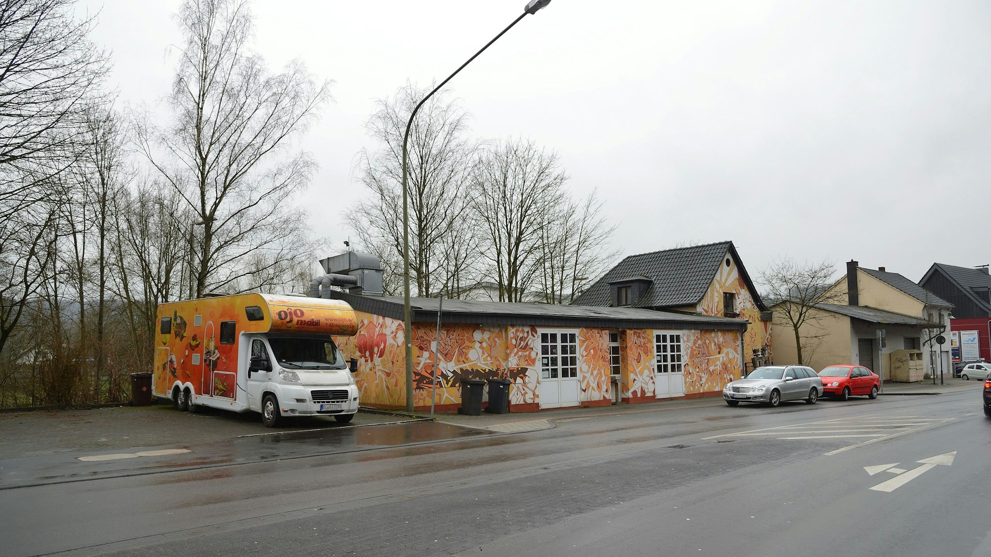 Ein gelb- und orangefarben angemaltes Haus steht an einer Hauptstraße, daneben befindet sich ein Wohnmobil in den selben Farben.