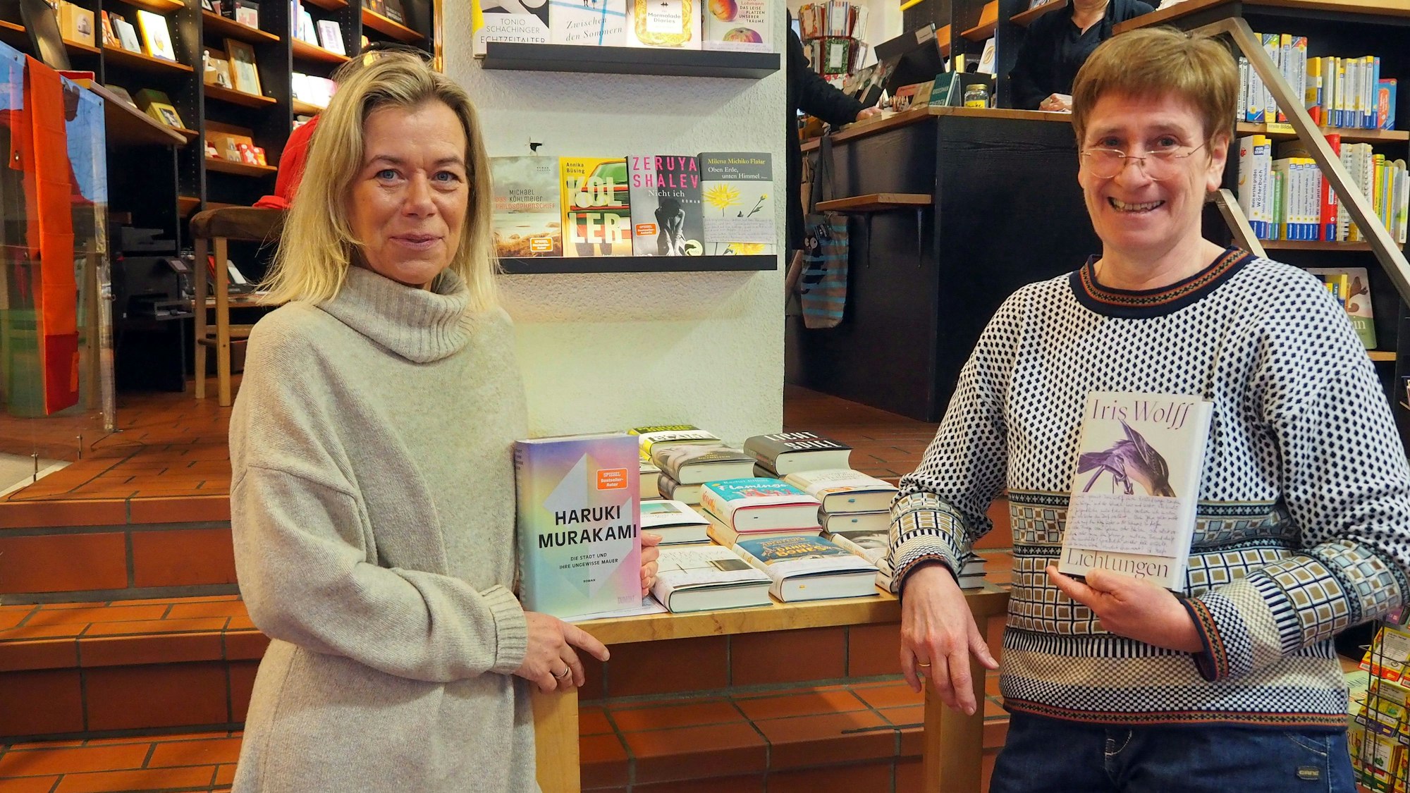 Zwei Frauen stehen in einer Buchhandlung. Jede hat ein Buch in der Hand.