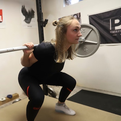 Beim Training stemmt Helena Decker Gewichte. Dabei geht sie in die Knie. Auf einem Poster im Hintergrund steht „Stay Metal“.