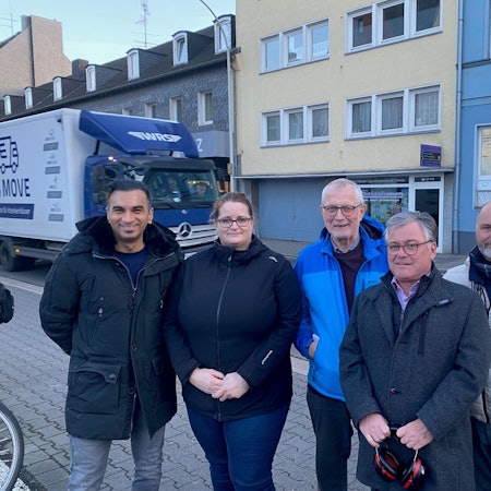 Zu sehen ist eine Gruppe von Bürgern vor der Heinrich-Esser-Straße, die gerade von einem Lkw befahren wird.