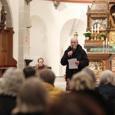 Ein Geistlicher in schwarzem Anorak steht in einer voll besetzten Kirche und spricht in ein Mikrofon, eine Frau sitzt daneben.