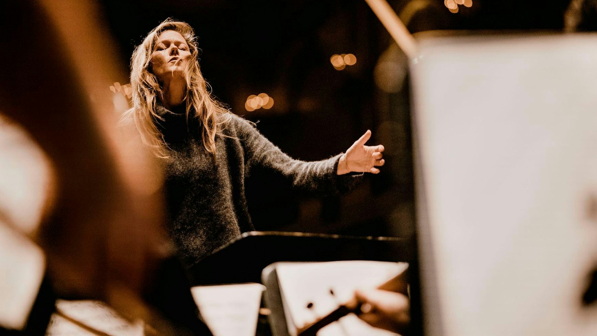 Das Bild zeigt Barbara Hannigan mit geschlossenen Augen beim Dirigieren. Im Vordergrund sind einige verschwommene Pulte mit Notenblättern zu sehen.