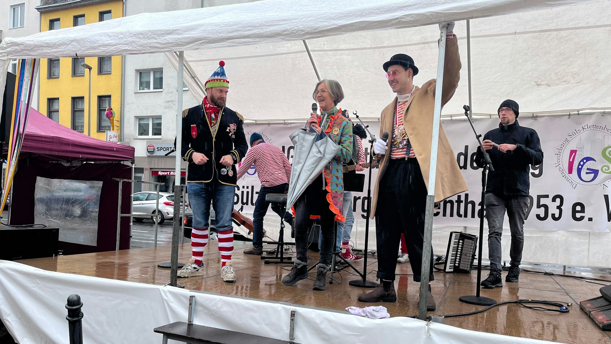 Auf einer Bühne stehen drei Männer, eine grauhaarige Dame spricht ins Mikrofon.