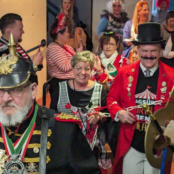 Eine kostümierte Frau führt den ebenfalls kostümierten Bürgermeister an der Kette durch einen gut gefüllten Saal.