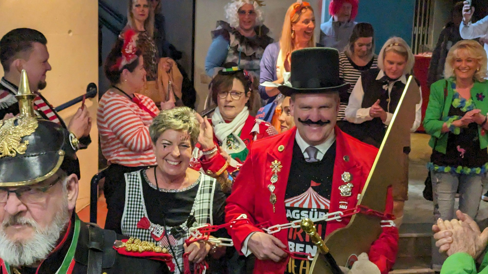 Eine kostümierte Frau führt den ebenfalls kostümierten Bürgermeister an der Kette durch einen gut gefüllten Saal.