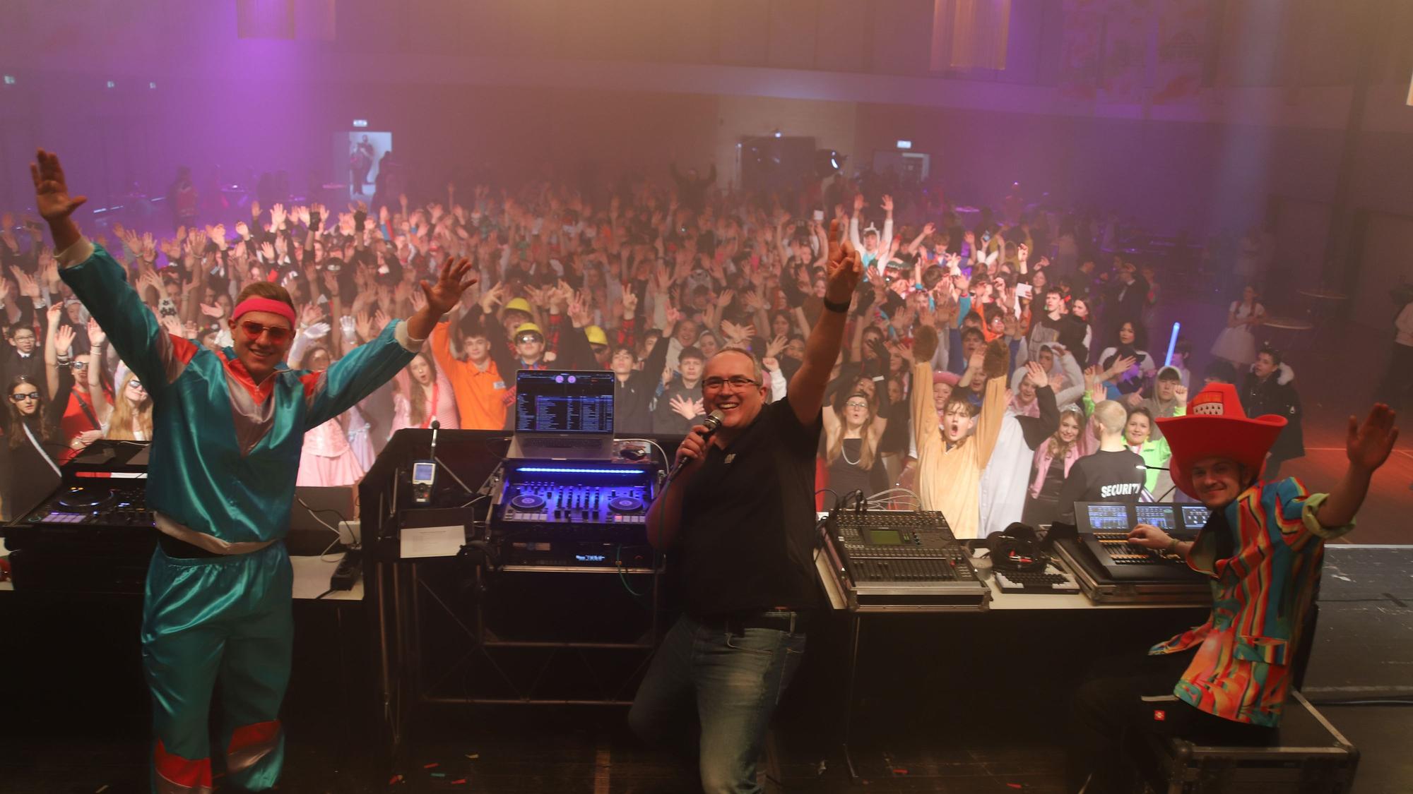 Blick von der Bühne in die Halle. Drei Männer stehen verkleidet auf der Bühne, im Hintergrund sind Jugendliche zu sehen. Alle Blicken in die Kamera.