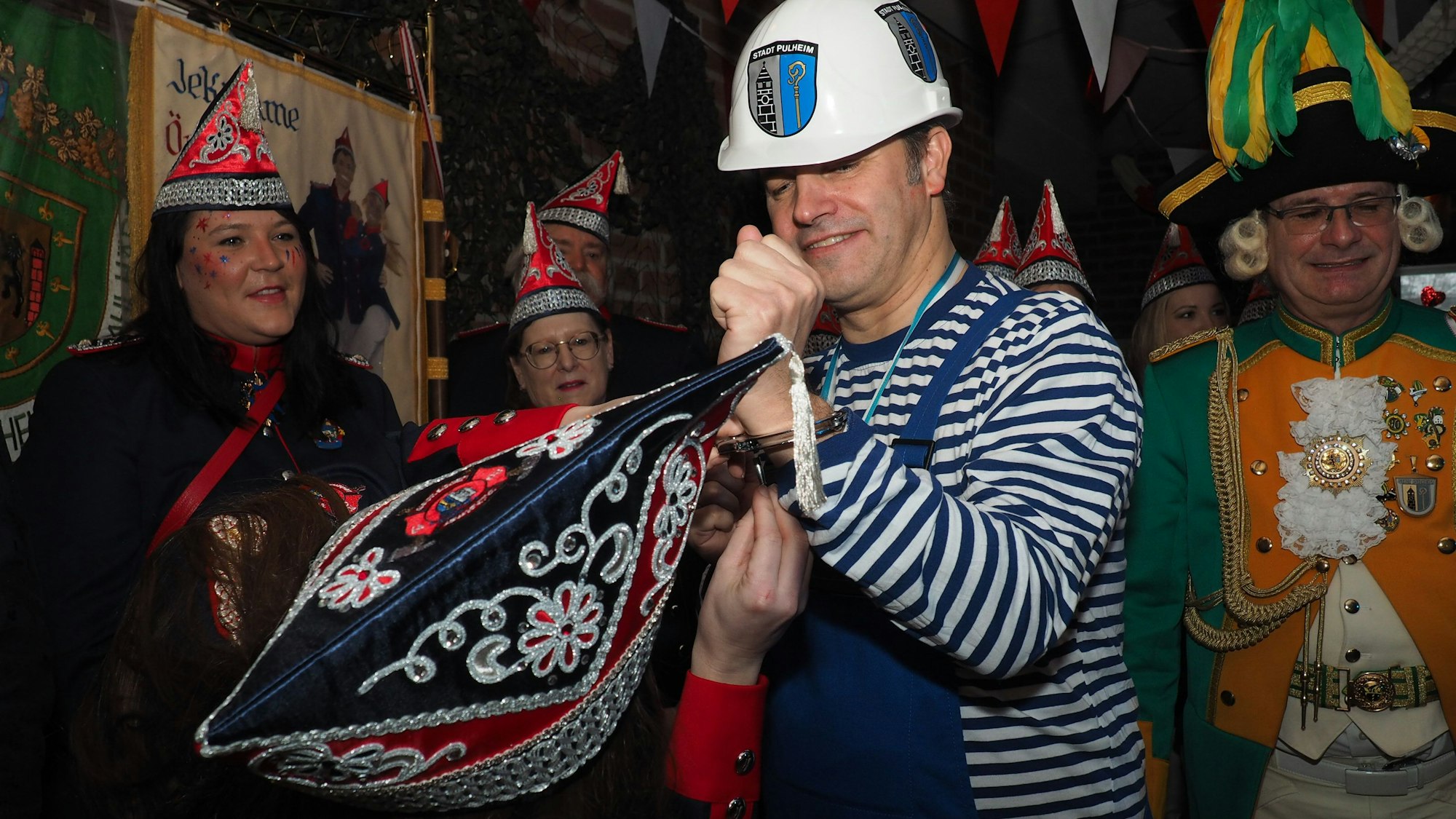 Pulheims Bürgermeister im Blaumann, darunter ein Ringelshirt in den Farben Blau und Weiß. Ein Mitglied der KG Löschgrenadiere versucht, die Handschellen zu lösen.