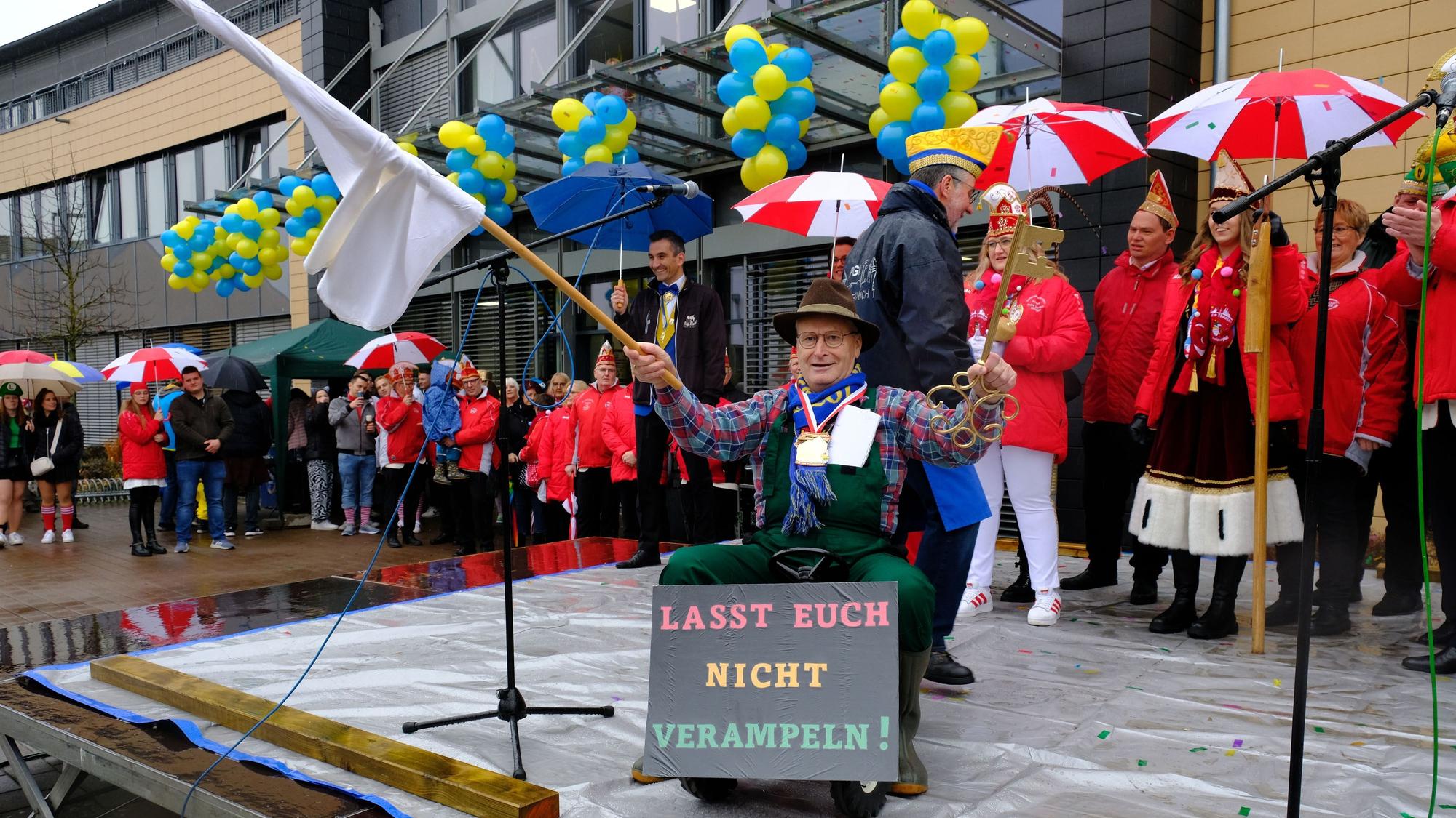 Bürgermeister Hans-Peter Schick sitzt auf einem Spielzeugtrecker und schwenkt die weiße Fahne. In der linken Hand hält er den Stadtschlüssel, den er an die Jecken abgibt.