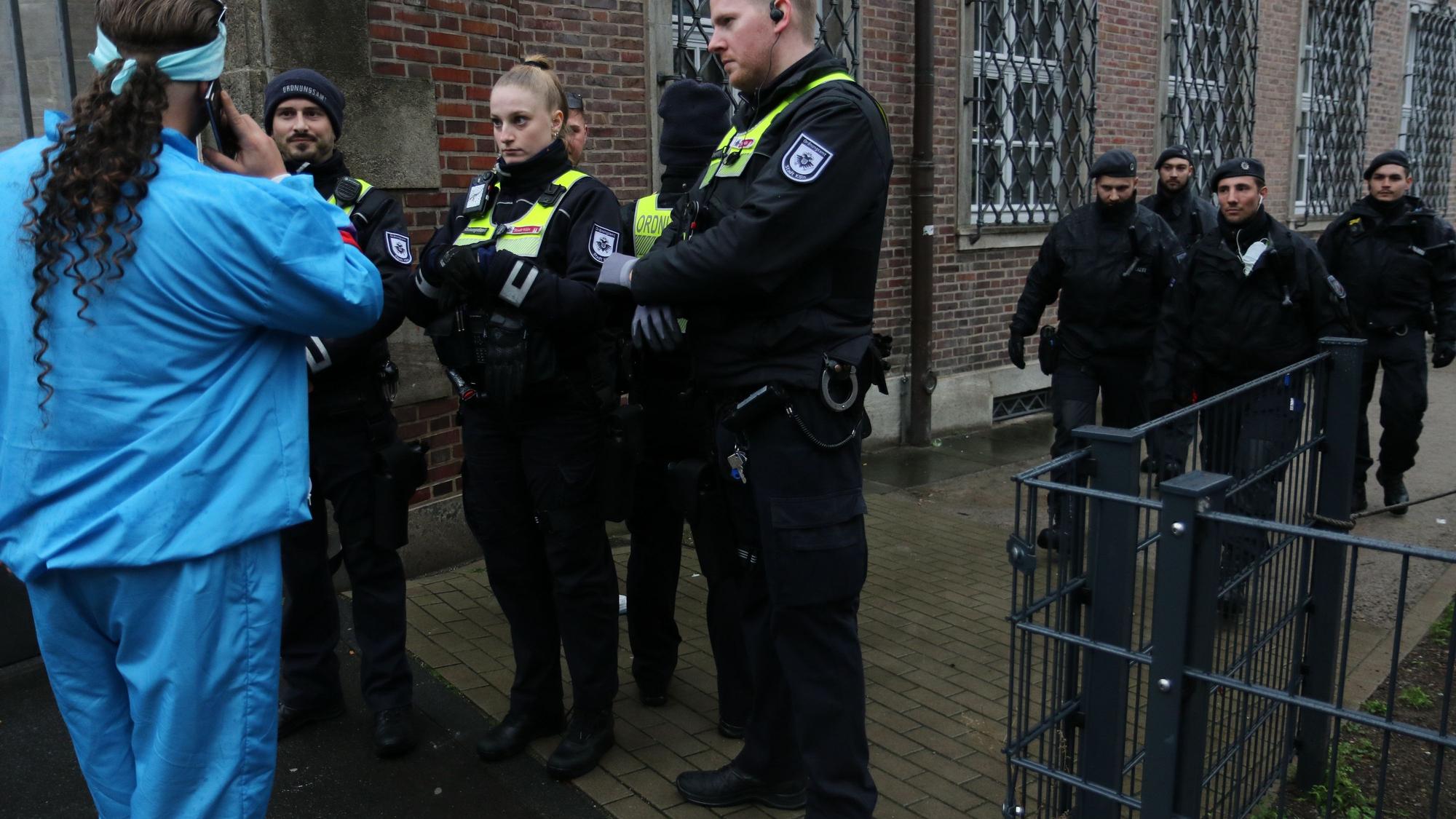 Mitarbeiter des Ordnungsamtes stehen vor einem verkleideten Menschen, im Hintergrund laufen Polizisten an ihnen vorbei.