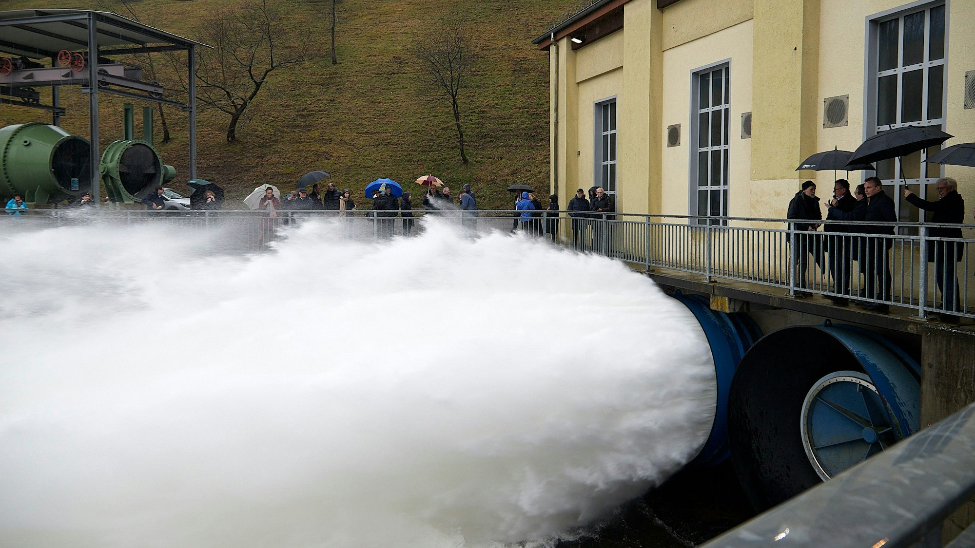 Aus großen Rohren unter einem gelben Gebäude schießt Wasser. Drumherum stehen Menschen mit Regenschirmen.
