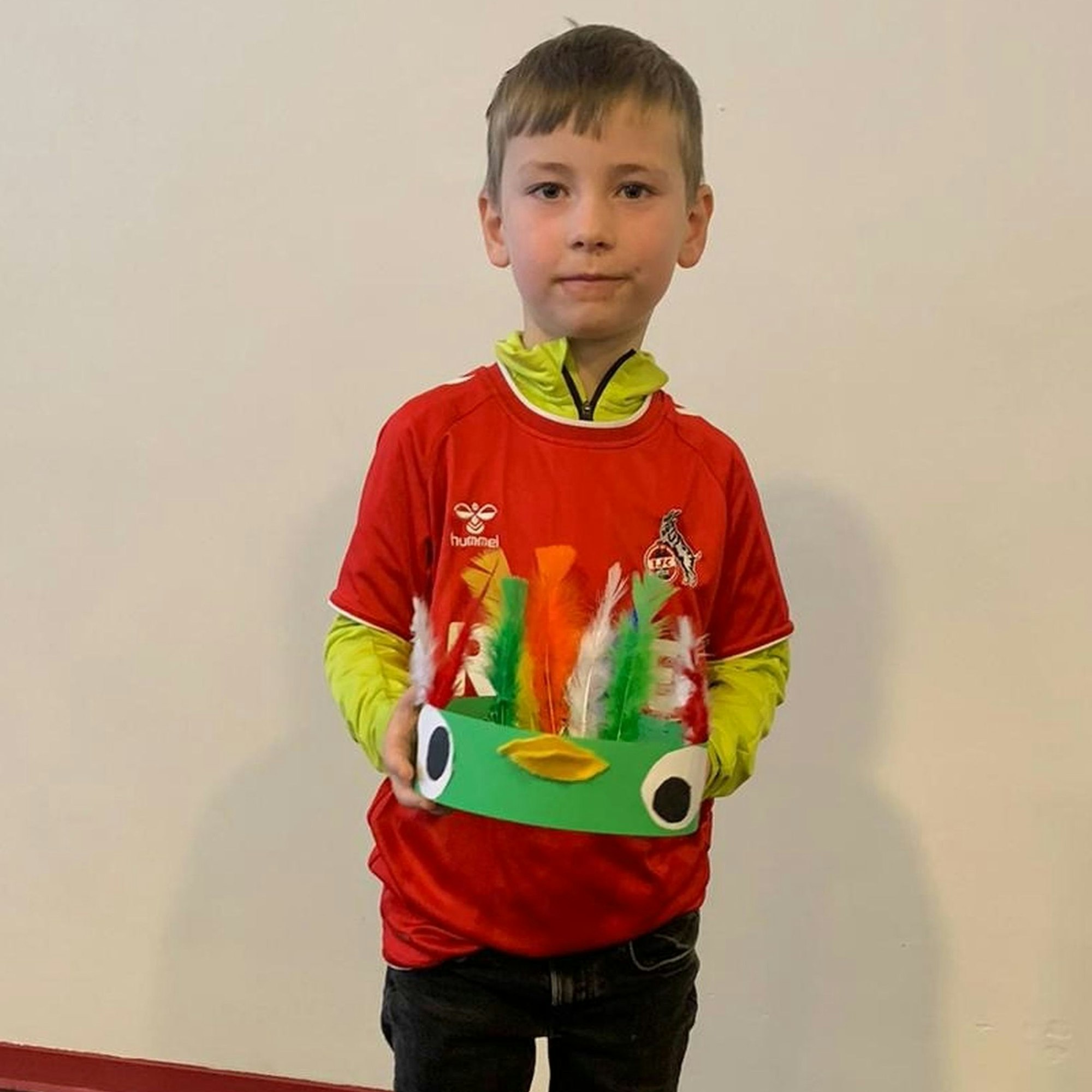 Julian Schmaul-Klaiber trägt ein Trikot des 1. FC Köln und hält eine gebastelte Krone in den Händen, die den Kopf eines grünen Papageis darstellt.