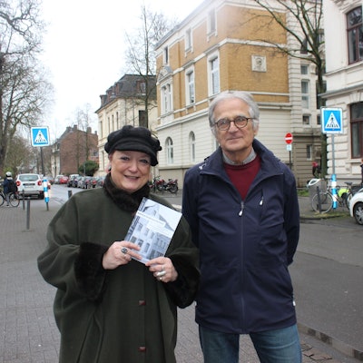 Eine Frau und ein Mann stehen auf einer Straße mit Altbauten. Sie hält ein Bändchen in der Hand.