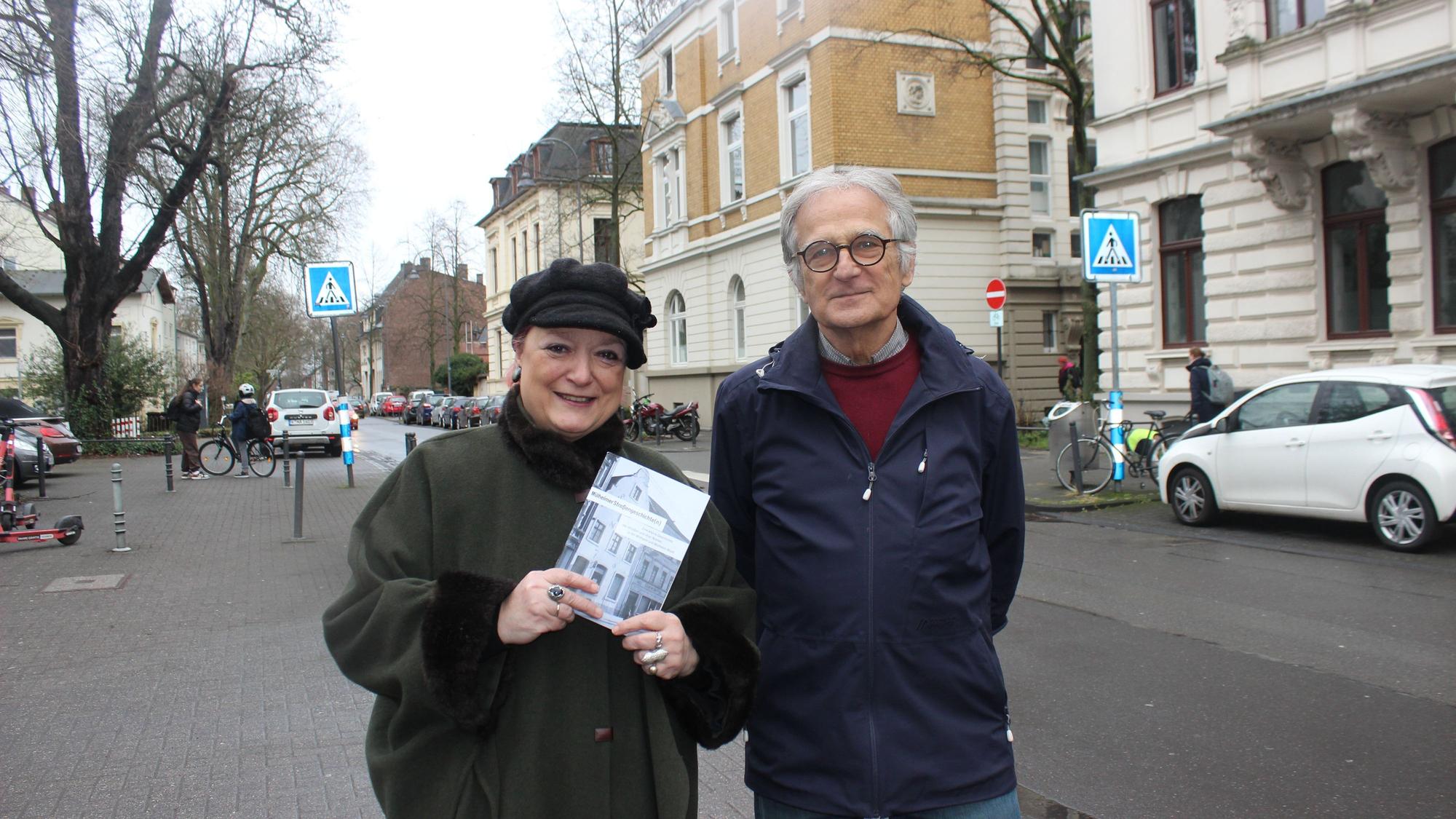 Eine Frau und ein Mann stehen auf einer Straße mit Altbauten. Sie hält ein Bändchen in der Hand.