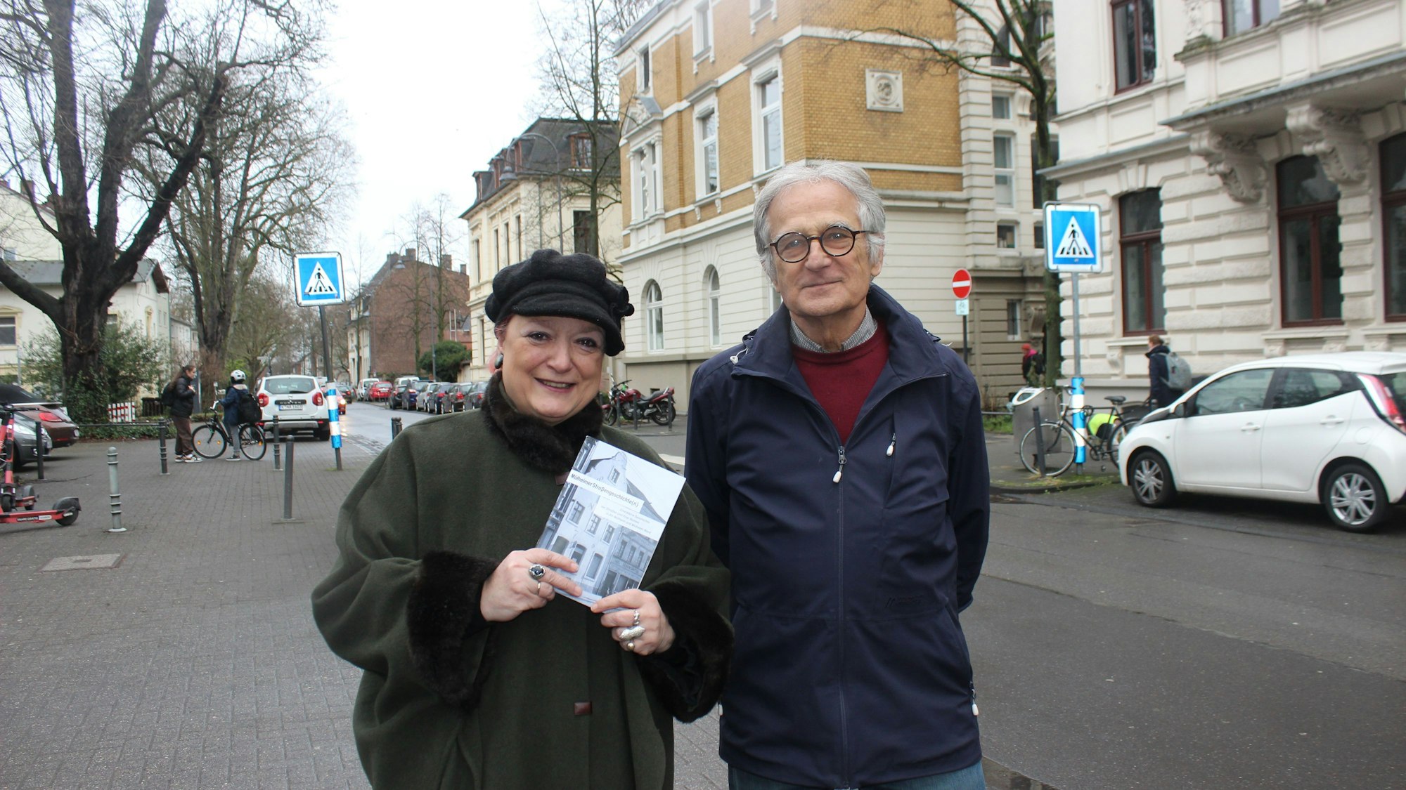 Eine Frau und ein Mann stehen auf einer Straße mit Altbauten. Sie hält ein Bändchen in der Hand.