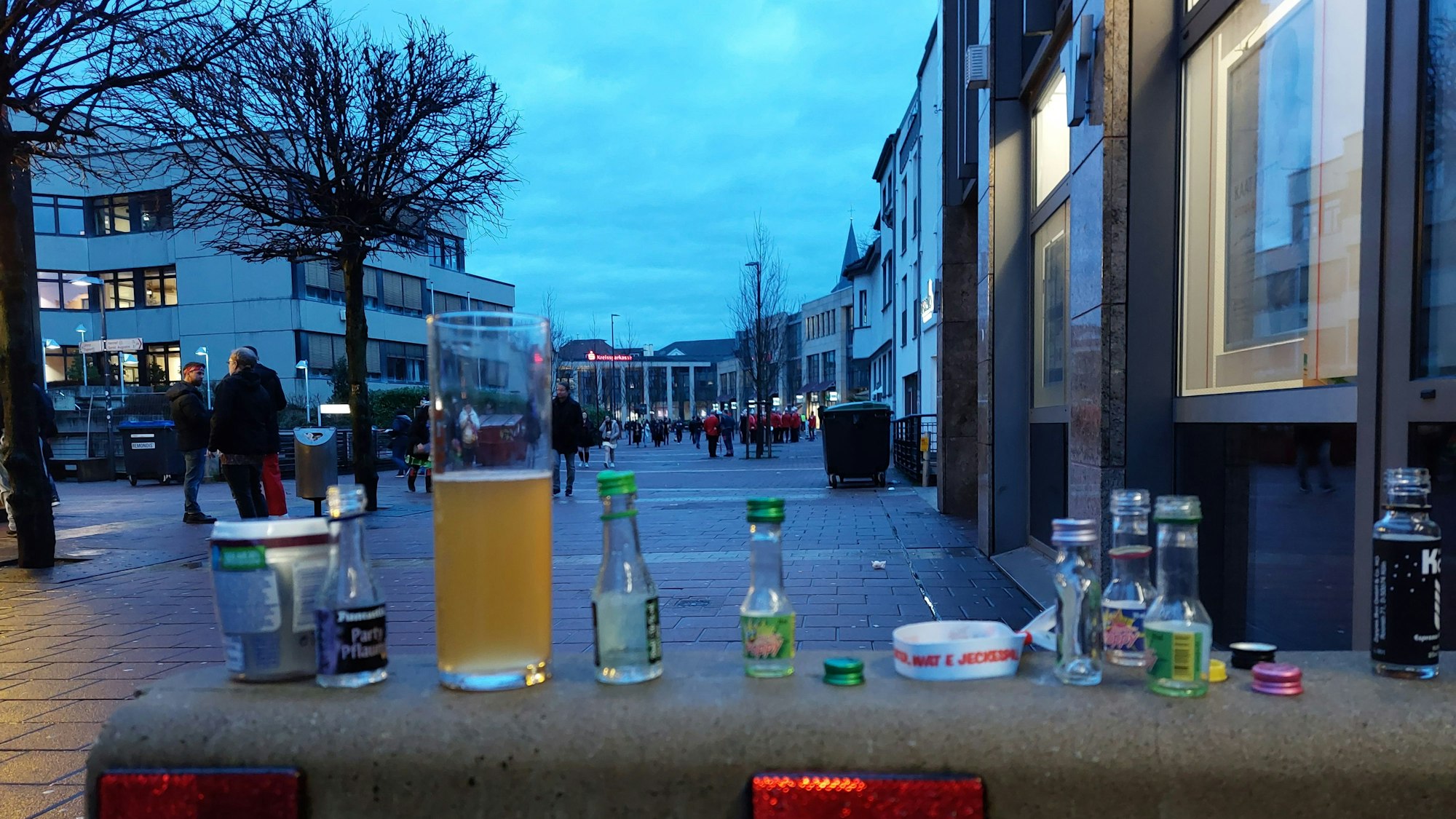 Leere Schnapsfläschchen und ein halbleeres Bierglas, bunte Flaschendeckel auf einer Betonsperre.