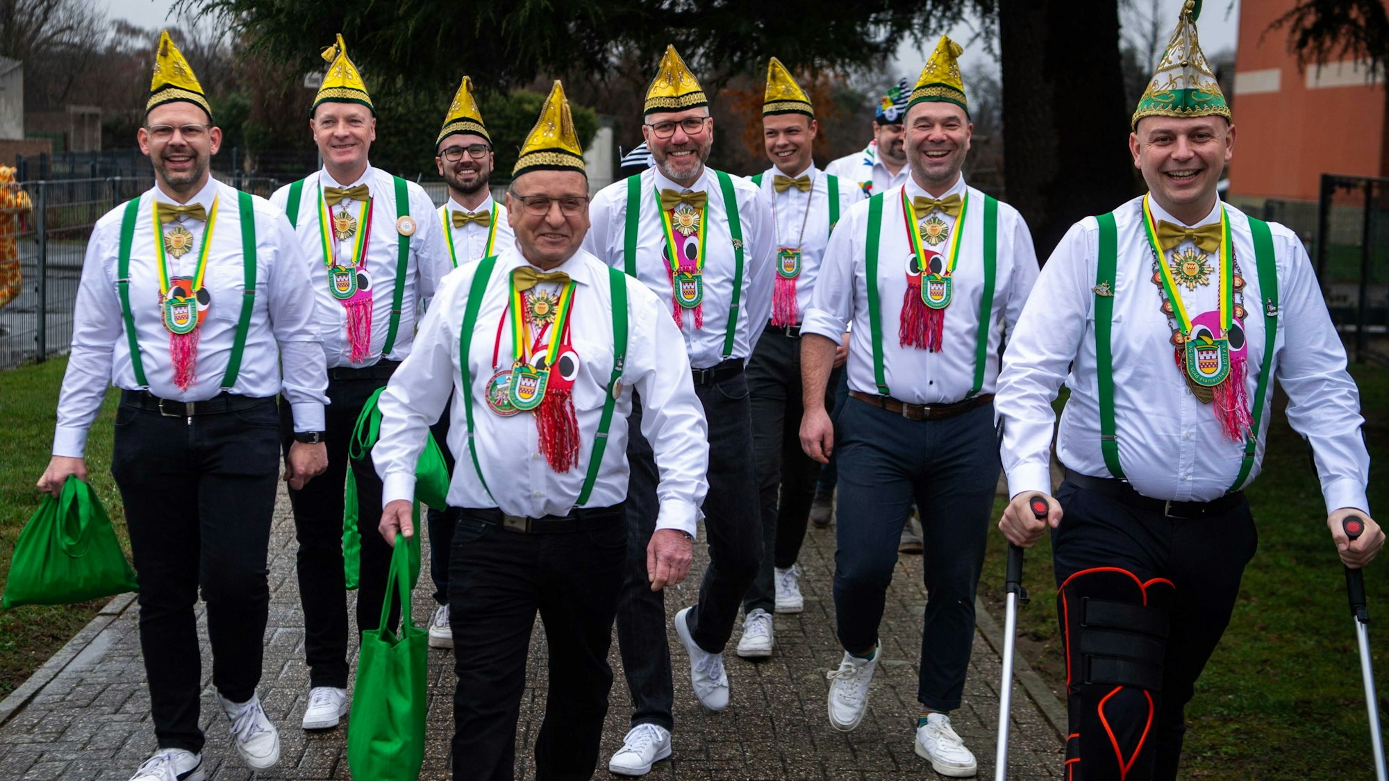 Neun Mitglieder der KG Flamersheim tragen weiße Hemden, grüne Hosenträger und goldene Narrenkappen.