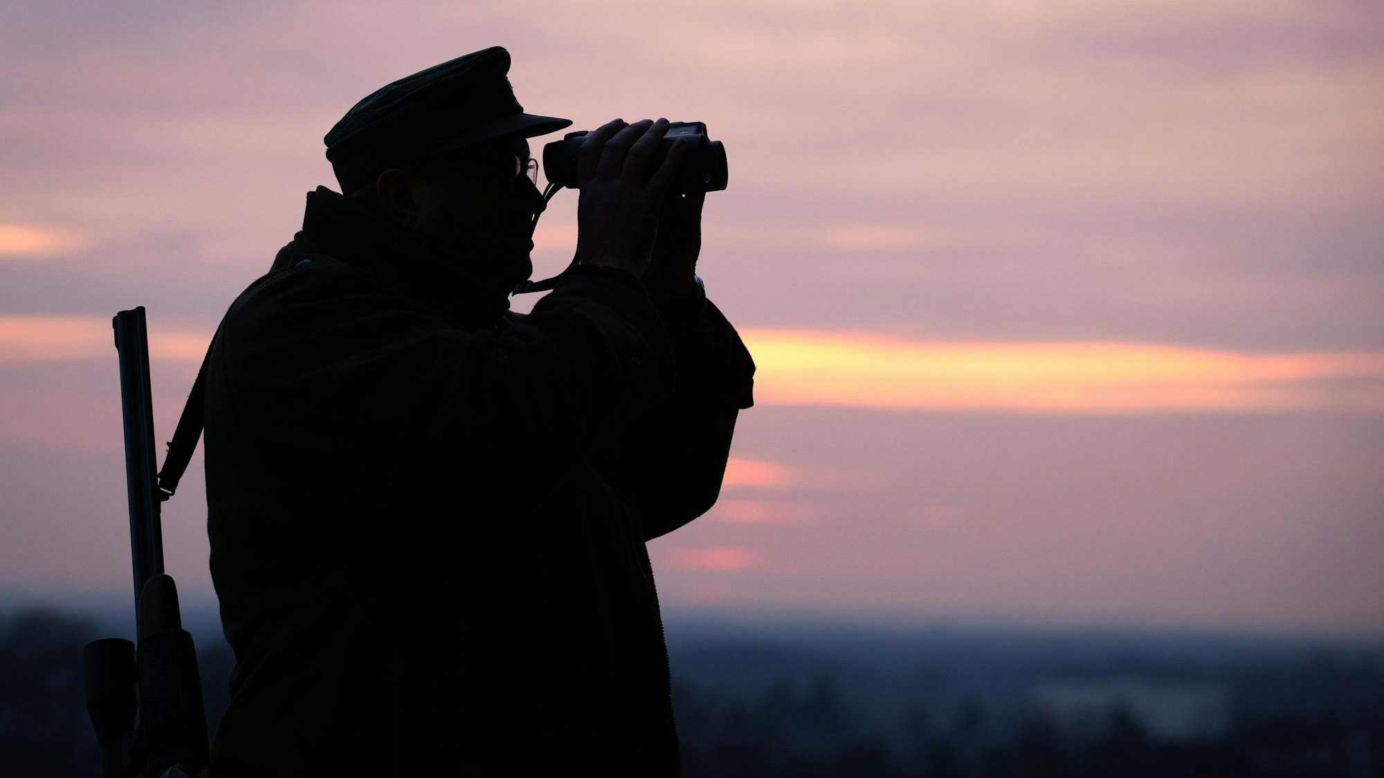 Ein Jäger blickt durch sein Fernglas in der Abenddämmerung.