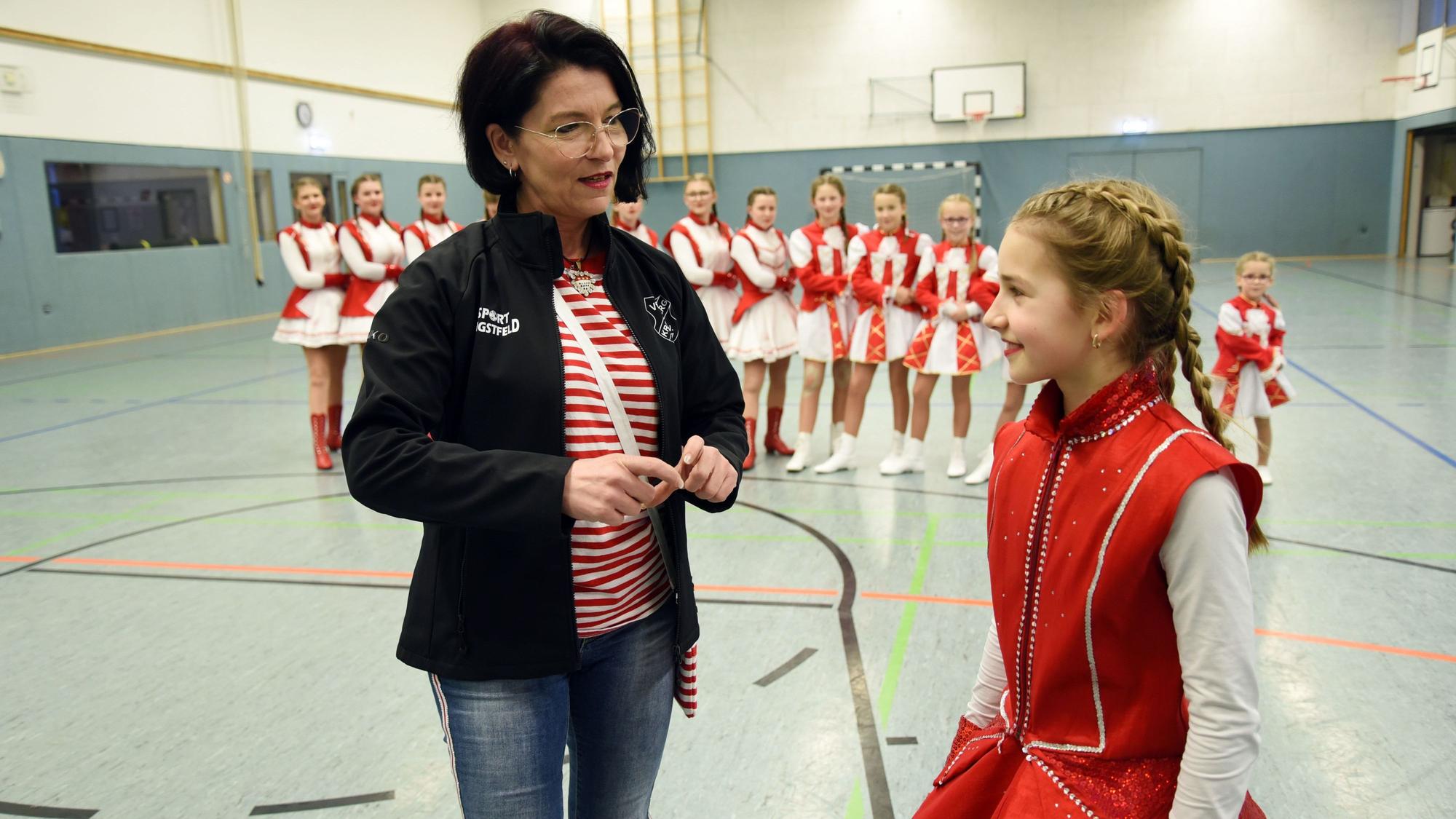 Trainerin Alex Jesinghaus steht mit dem Mariechen Merle Irle zusammen und spricht mit ihr. Im Hintergrund stehen die Mädchen der Tanzgruppe in einer Reihe.