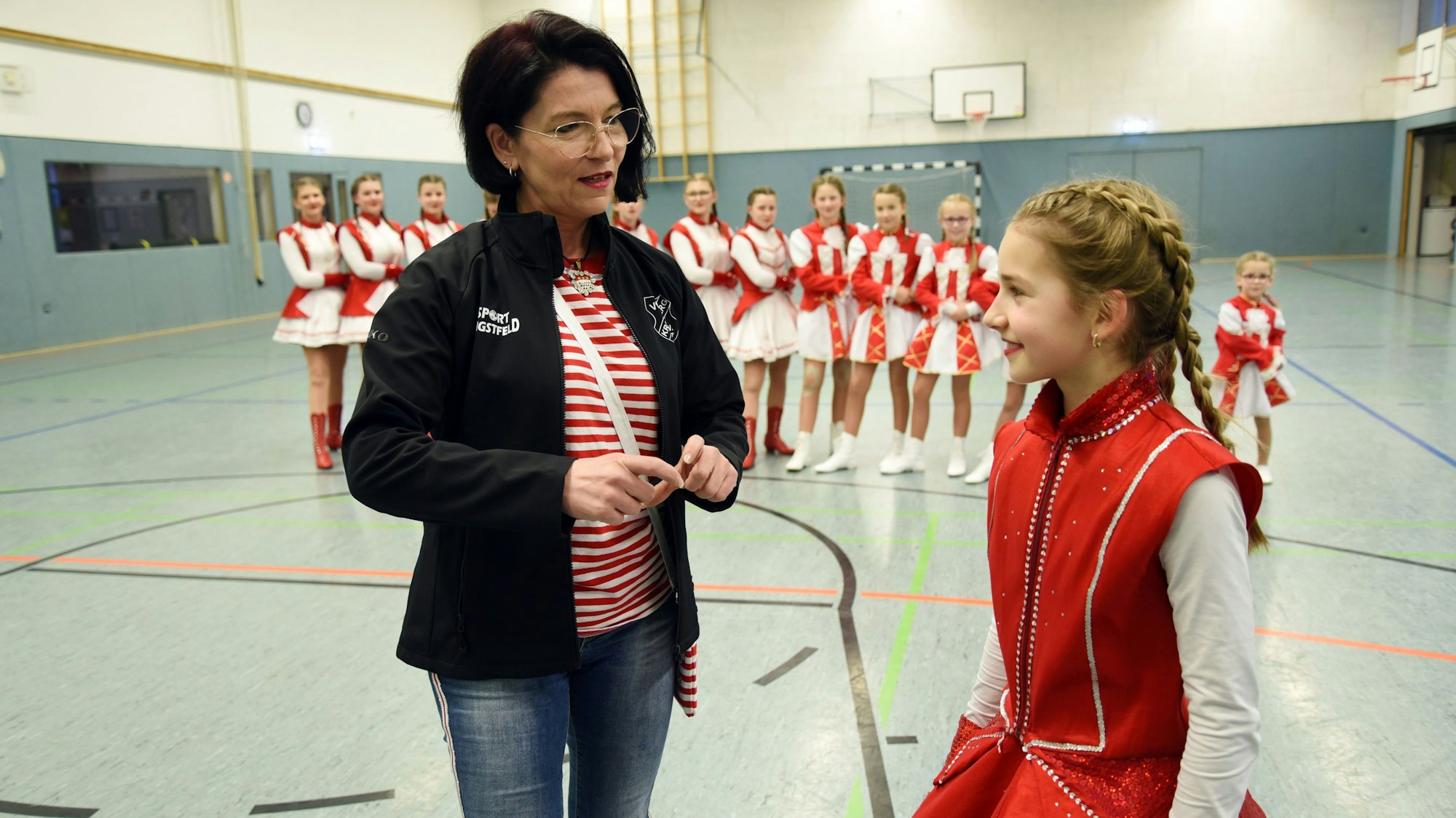 Trainerin Alex Jesinghaus steht mit dem Mariechen Merle Irle zusammen und spricht mit ihr. Im Hintergrund stehen die Mädchen der Tanzgruppe in einer Reihe.