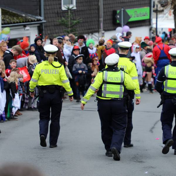 Polizisten gehen auf der Kürtener Bergstraße, auf der der Karnevalszug herannaht. Am Rand stehen kostümierte Jecke.