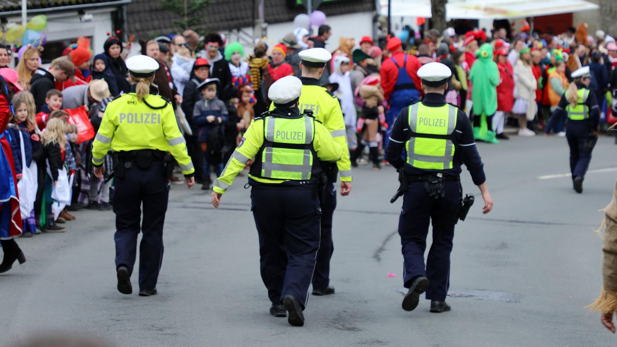Polizisten gehen auf der Kürtener Bergstraße, auf der der Karnevalszug herannaht. Am Rand stehen kostümierte Jecke.