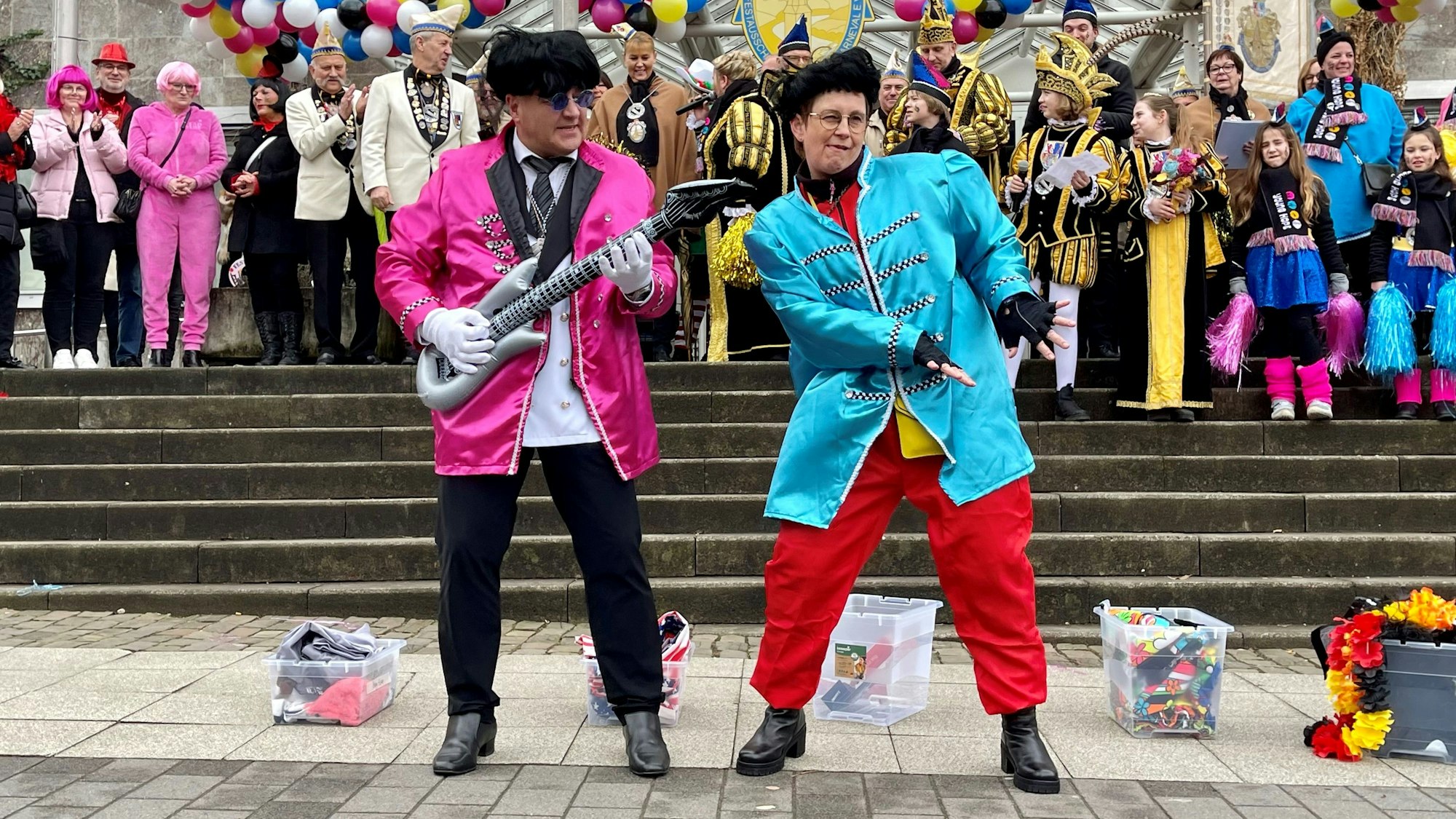 Ein Mann mit einer aufblasbaren Gitarre und eine Frau, beide in Pilzkopf-Perücken und bunten Jacken rocken vor der Rathaustreppe ab,