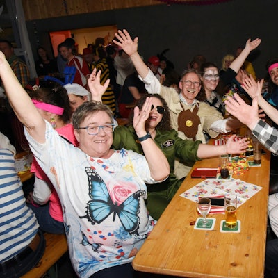 Mehrere verkleidete Leute sitzen gemeinsamen an einem Tisch, schauen in die Kamera und reißen jubelnd die Hände in die Luft.