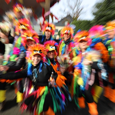 Menschen in Papageienkostümen stehen als Gruppe zusammen. Die Kamera zoomt schnell in das Bild hinein, dadurch kommt es zu Wischeffekten.