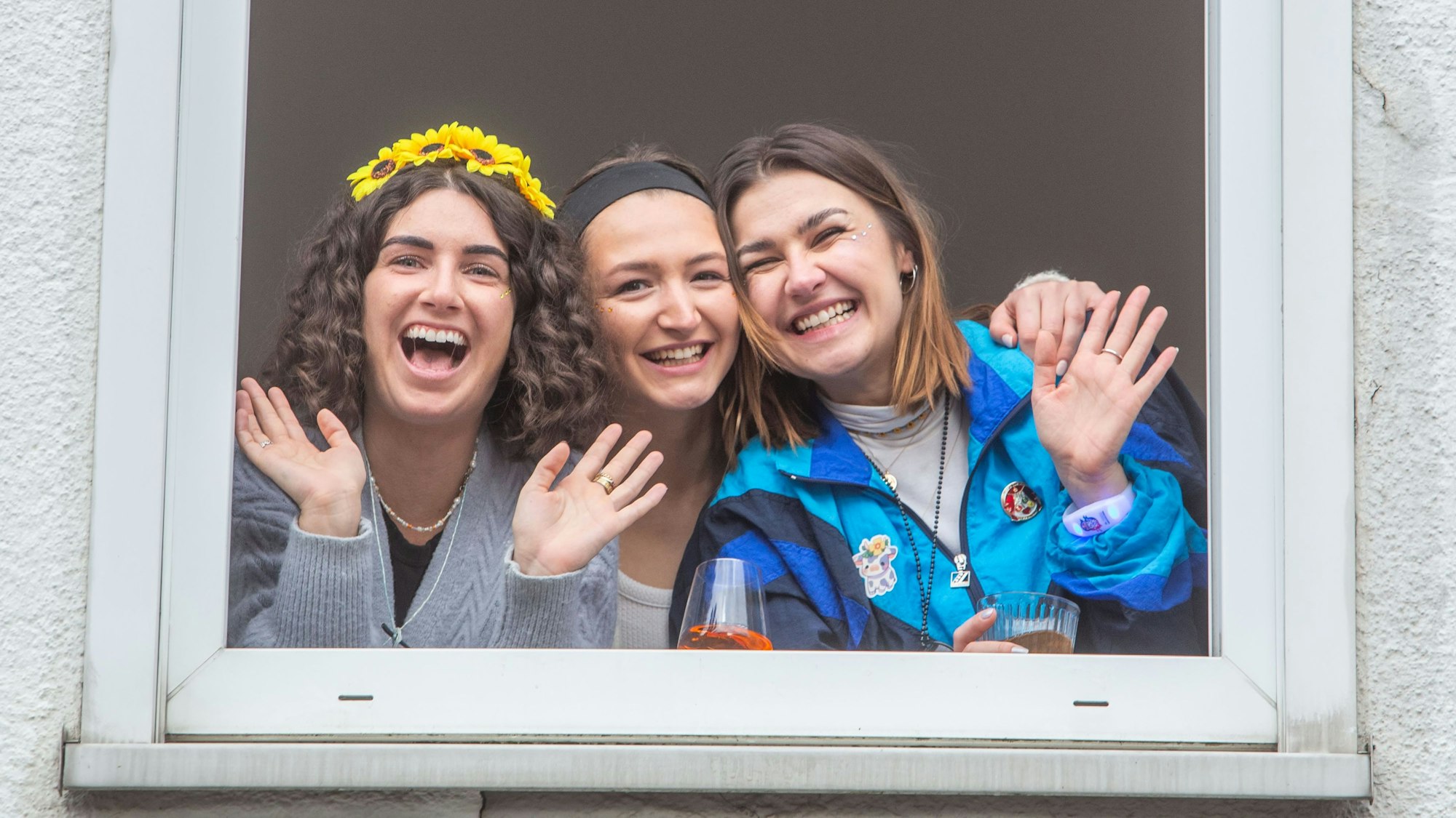 Drei verleidete junge Frauen winken aus einem Fenster.