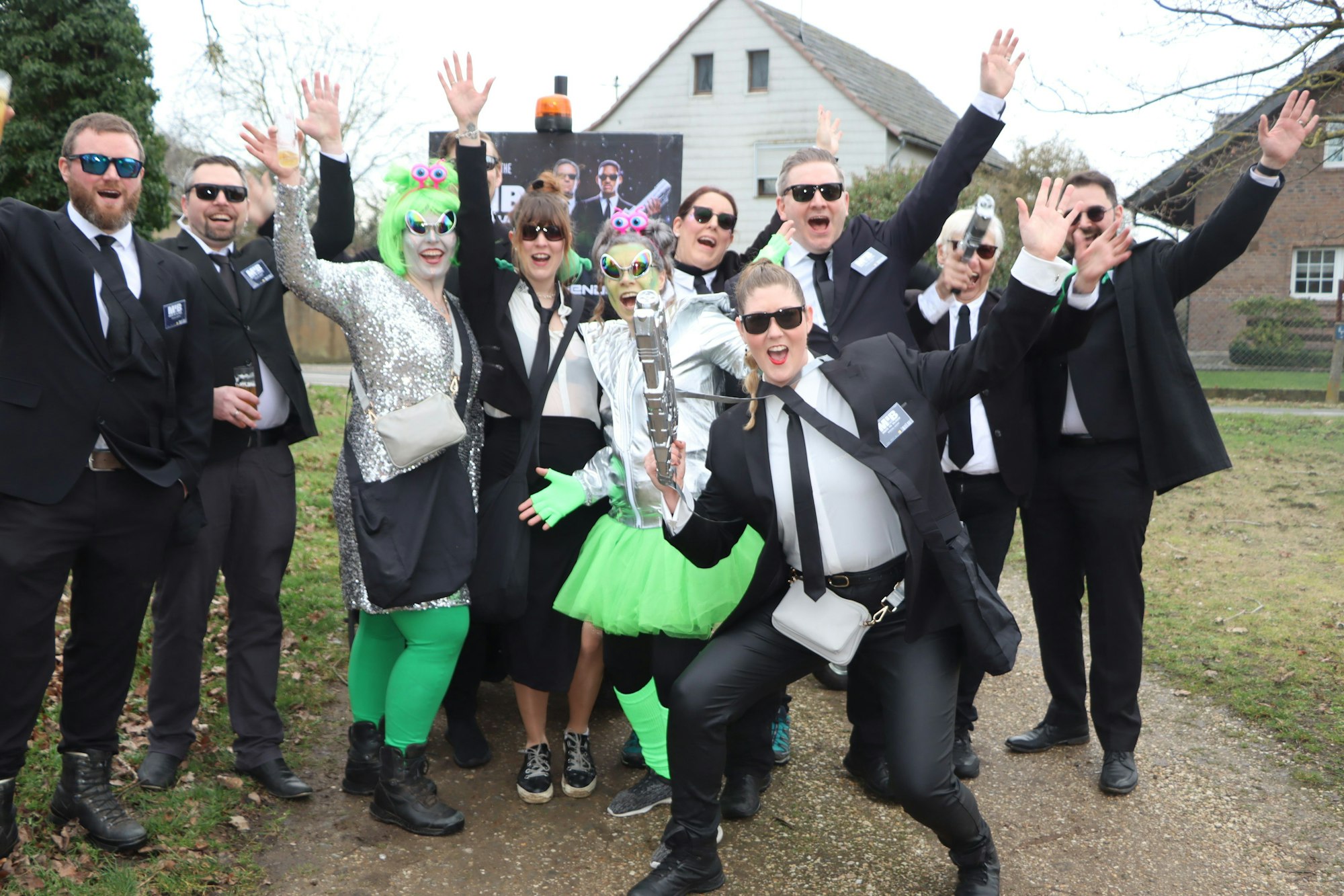 Männer in dunklen Anzügen und mit Sonnenbrillen gruppieren sich um weibliche Aliens.
