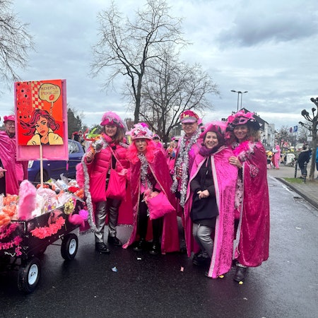 Eine Gruppe pink gekleideter Jecken mit einem Kamellewagen steht auf der Straße.