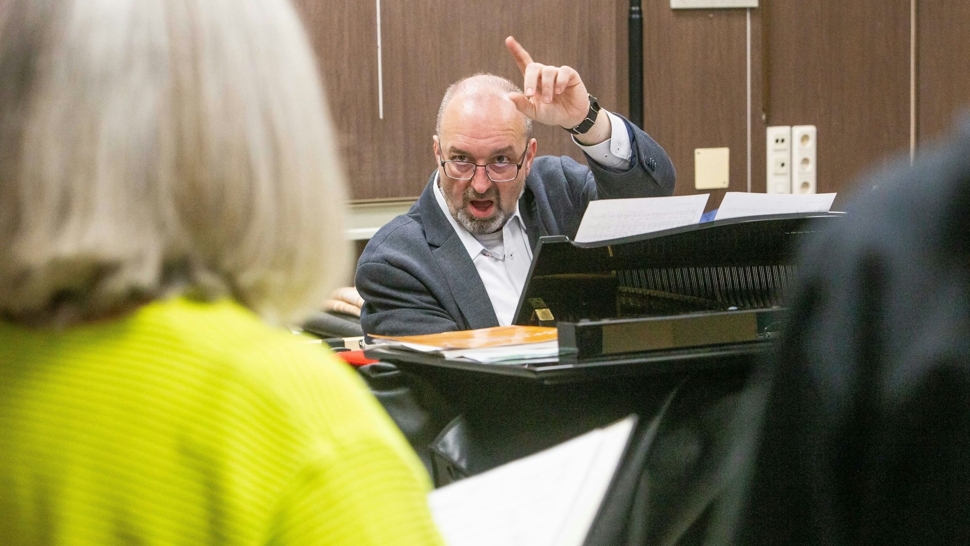 Ein Dirigent sitzt an einem Flügel und gibt einer Sängerin mit erhobenen Zeigefinger den Ton an.