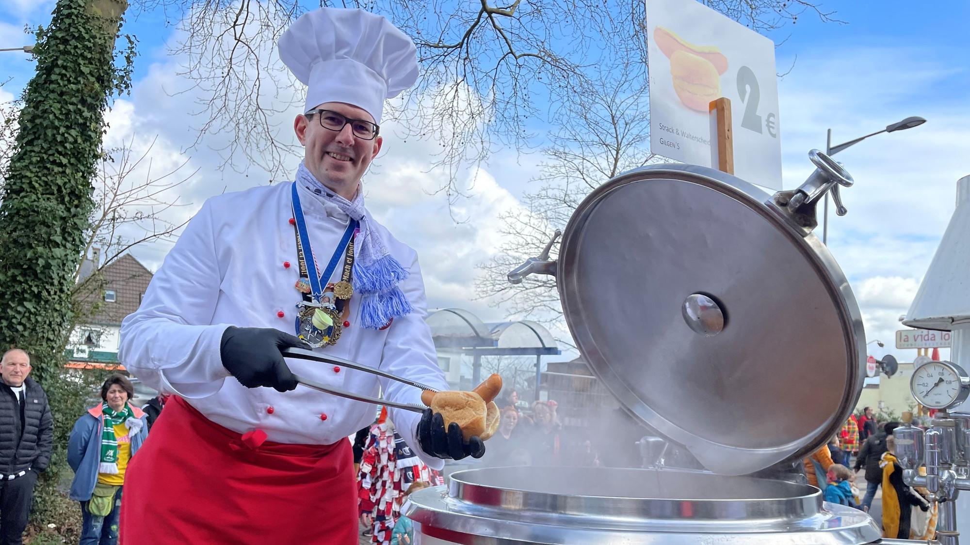 Ein als Koch verkleideter Mann legt mit einer Zange ein Knackwürstchen in ein aufgeschnittenes Brötchen.